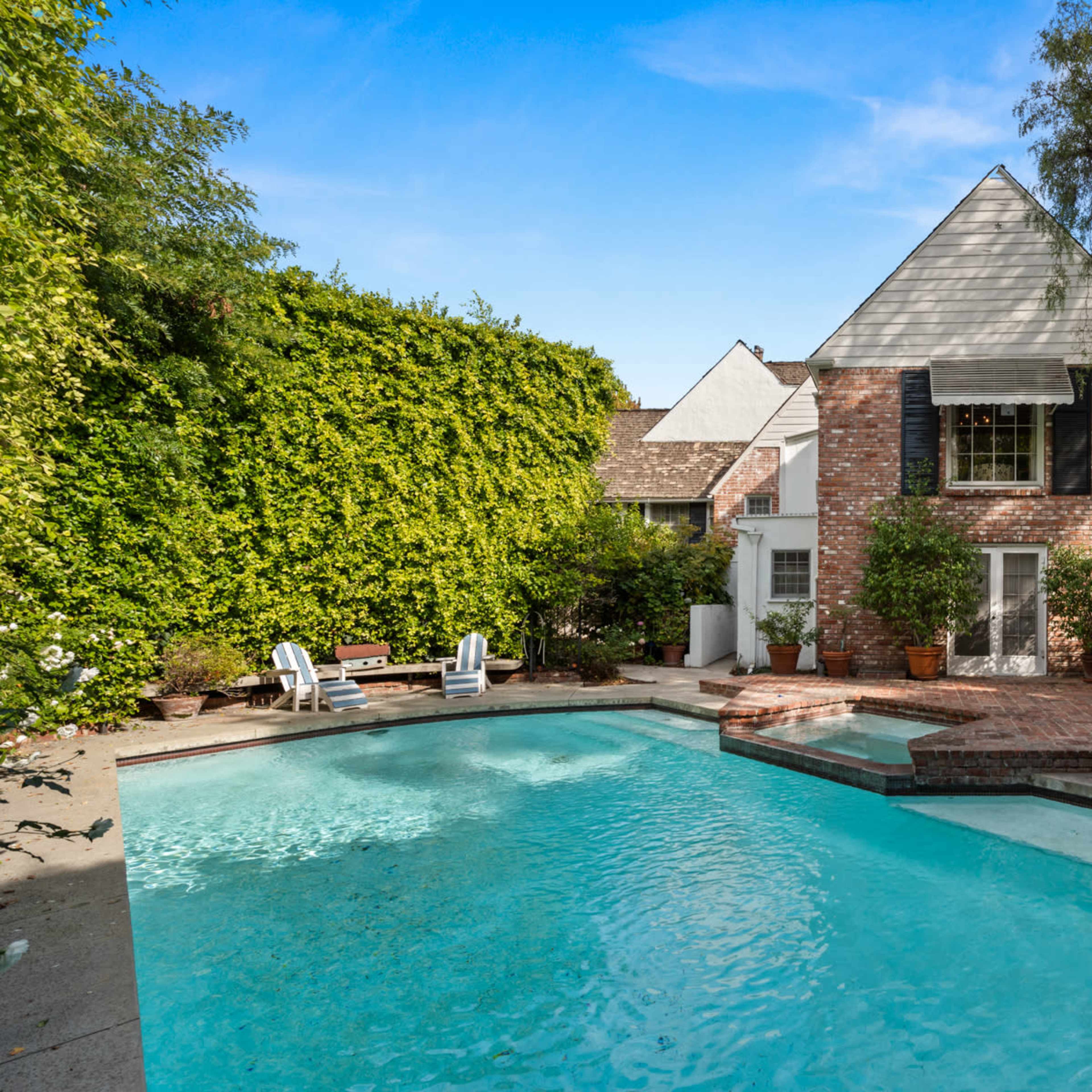 A backyard with a swimming pool, surrounded by greenery and a brick patio adjacent to a two-story house.
