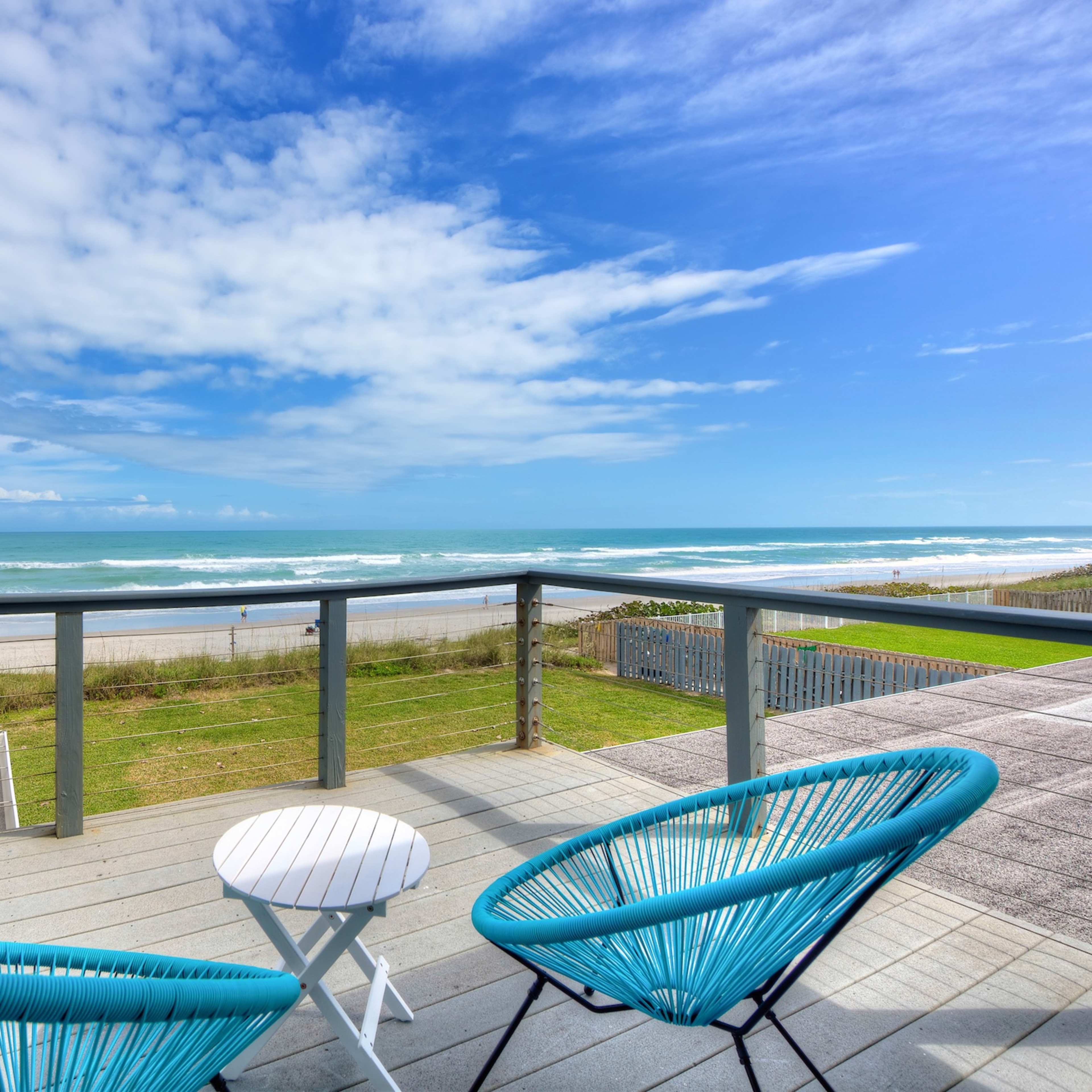 A balcony with two blue chairs overlooks a sandy beach and the ocean under a partly cloudy sky.