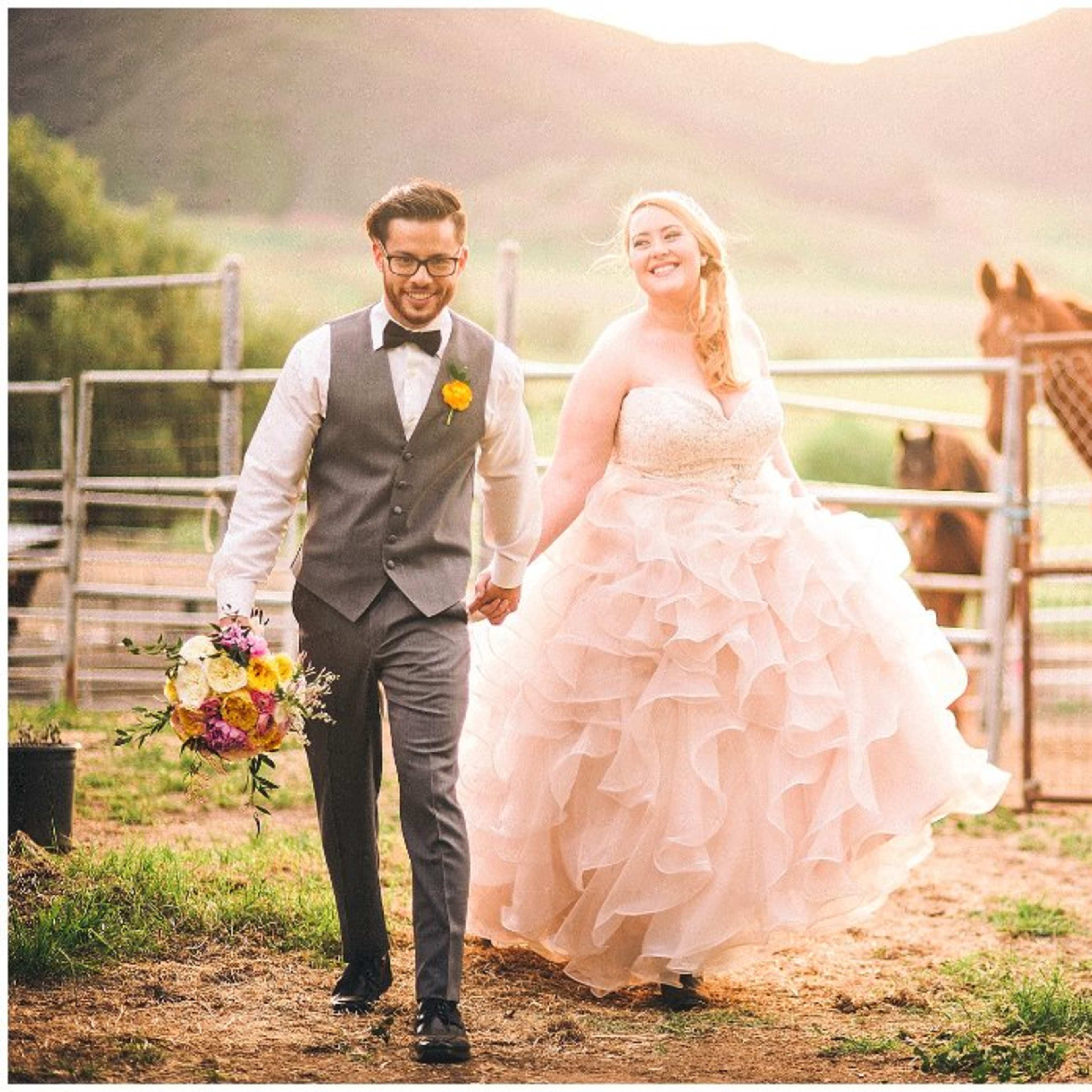A couple strolls hand in hand near a horse corral, with a scenic landscape in the background, while the bride holds a bouquet.
