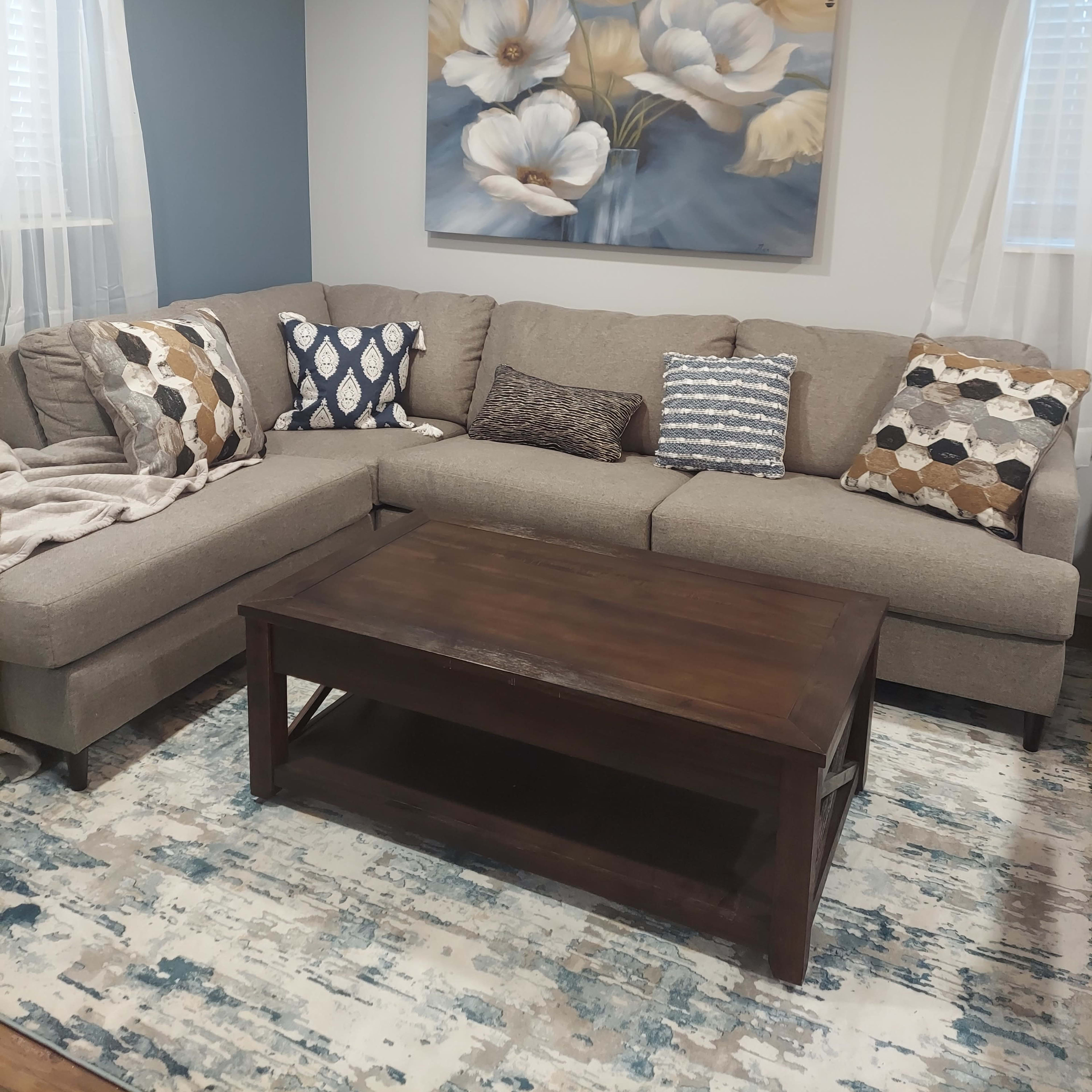 A living room with a sectional sofa, a wooden coffee table, and a floral painting on the wall.