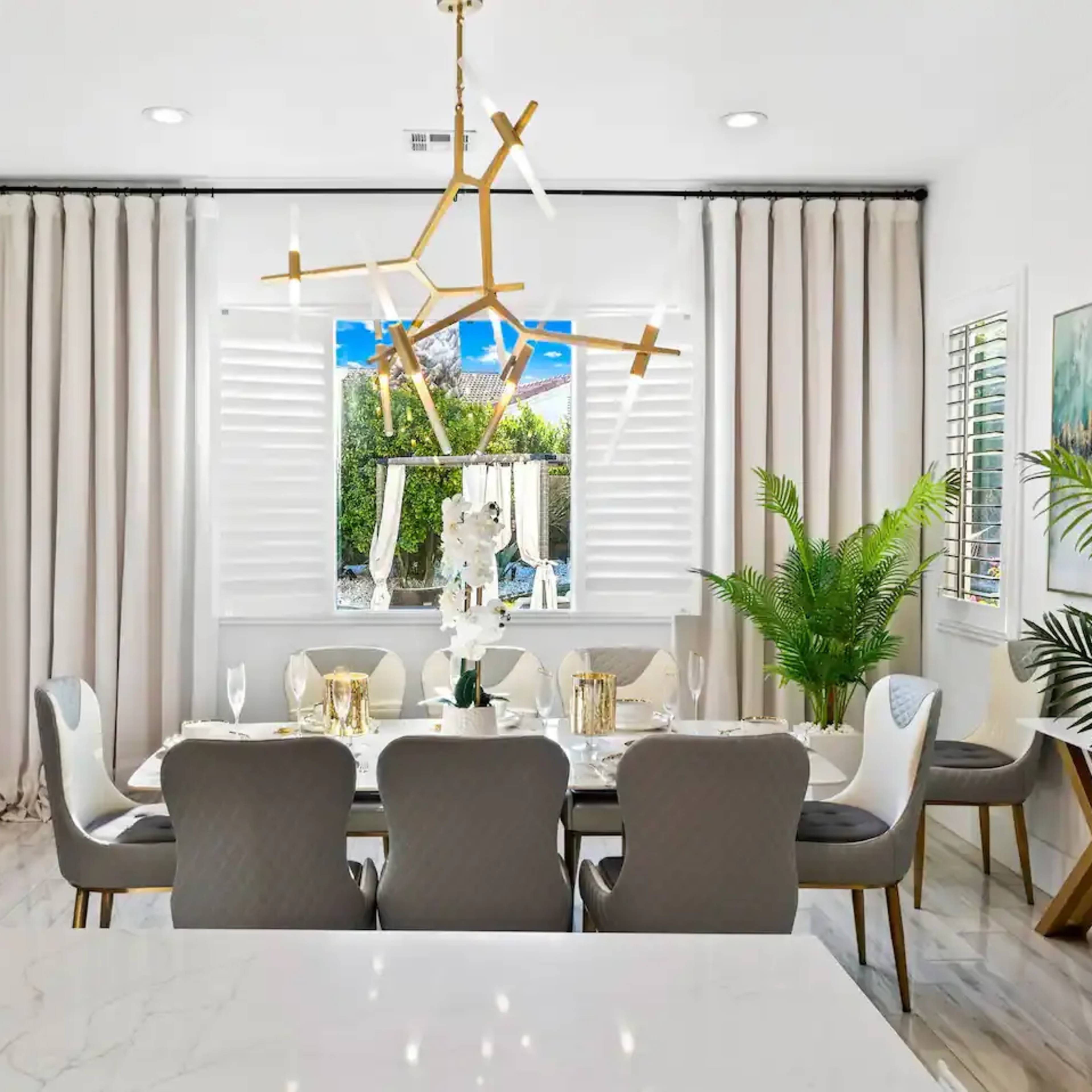 A dining room features a large table set for meals, surrounded by chairs, with a modern light fixture and potted plants near the windows.