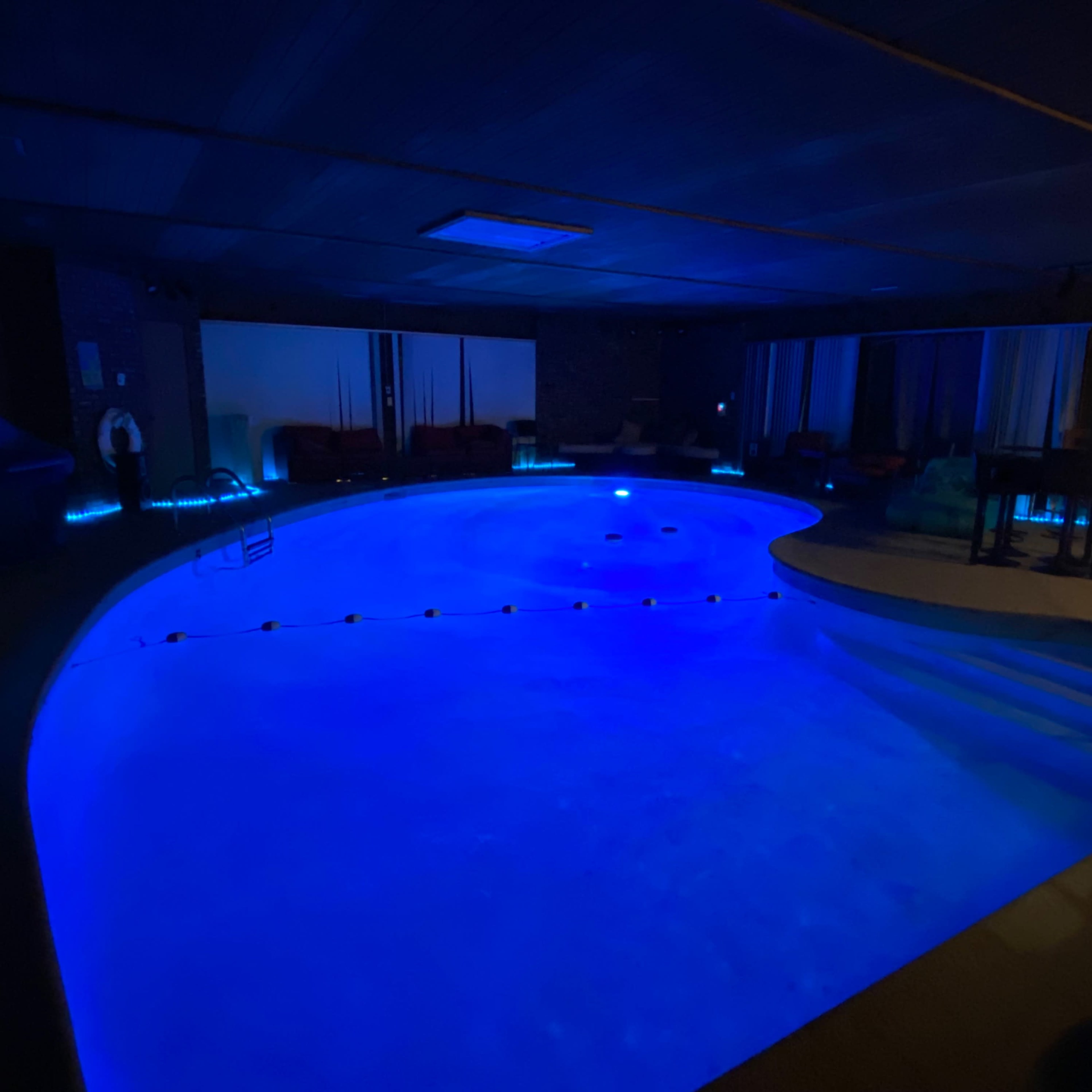 A dimly lit indoor swimming pool with blue lighting and lounge areas in the background.