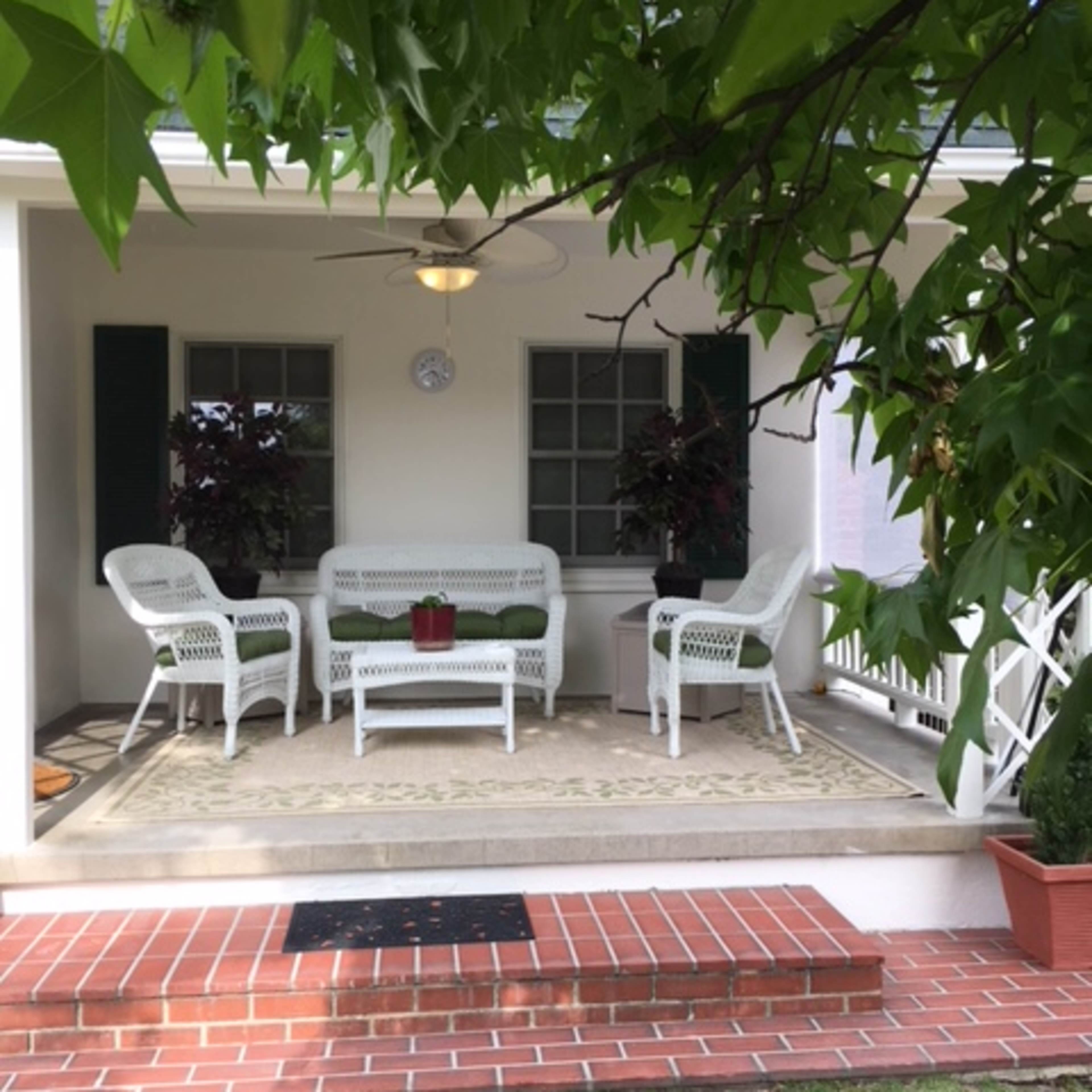 A porch with white wicker furniture arranged on a concrete floor, surrounded by green plants and a decorative rug.