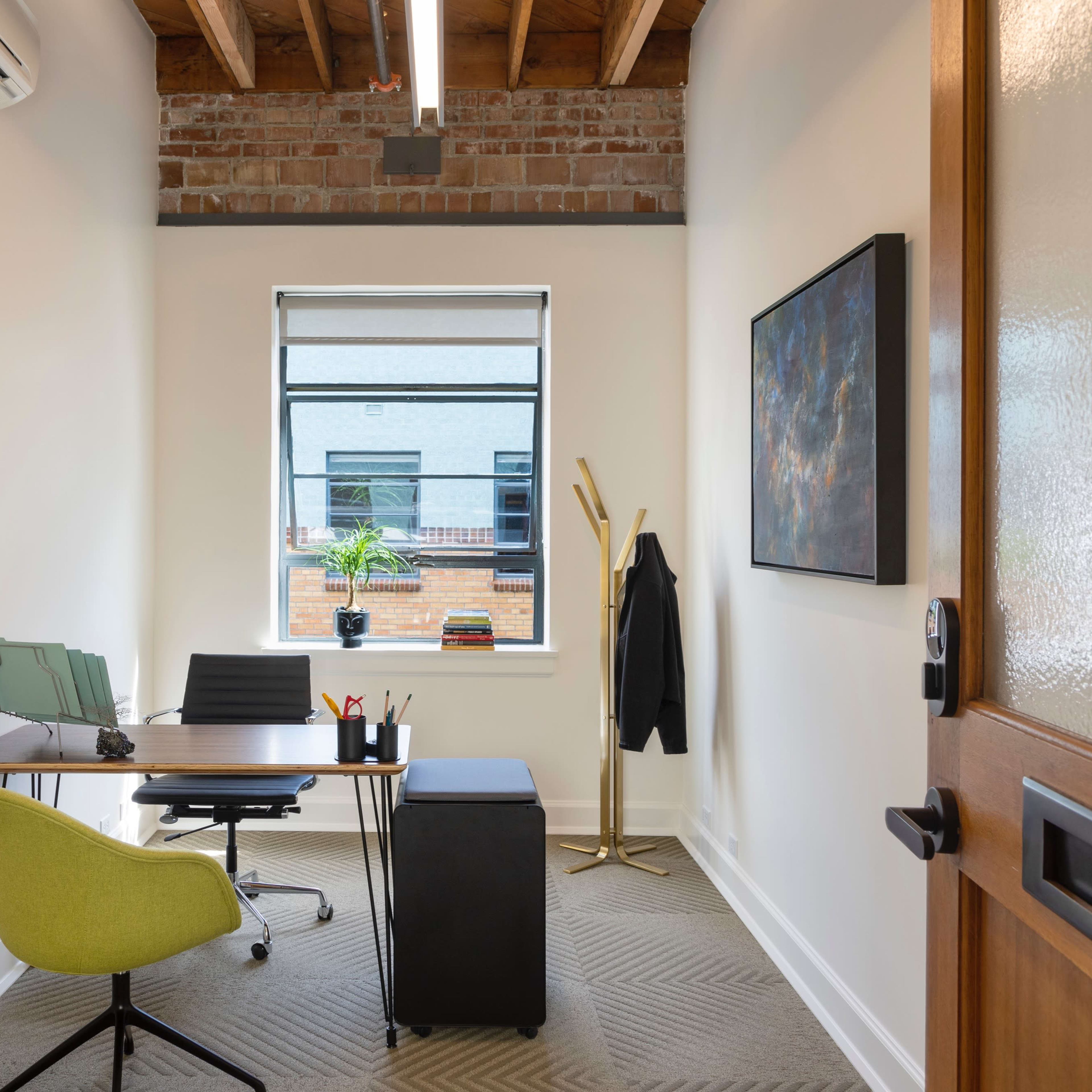 A small office featuring a desk with a green chair, a window with a view of a brick building, and a coat rack in the corner.
