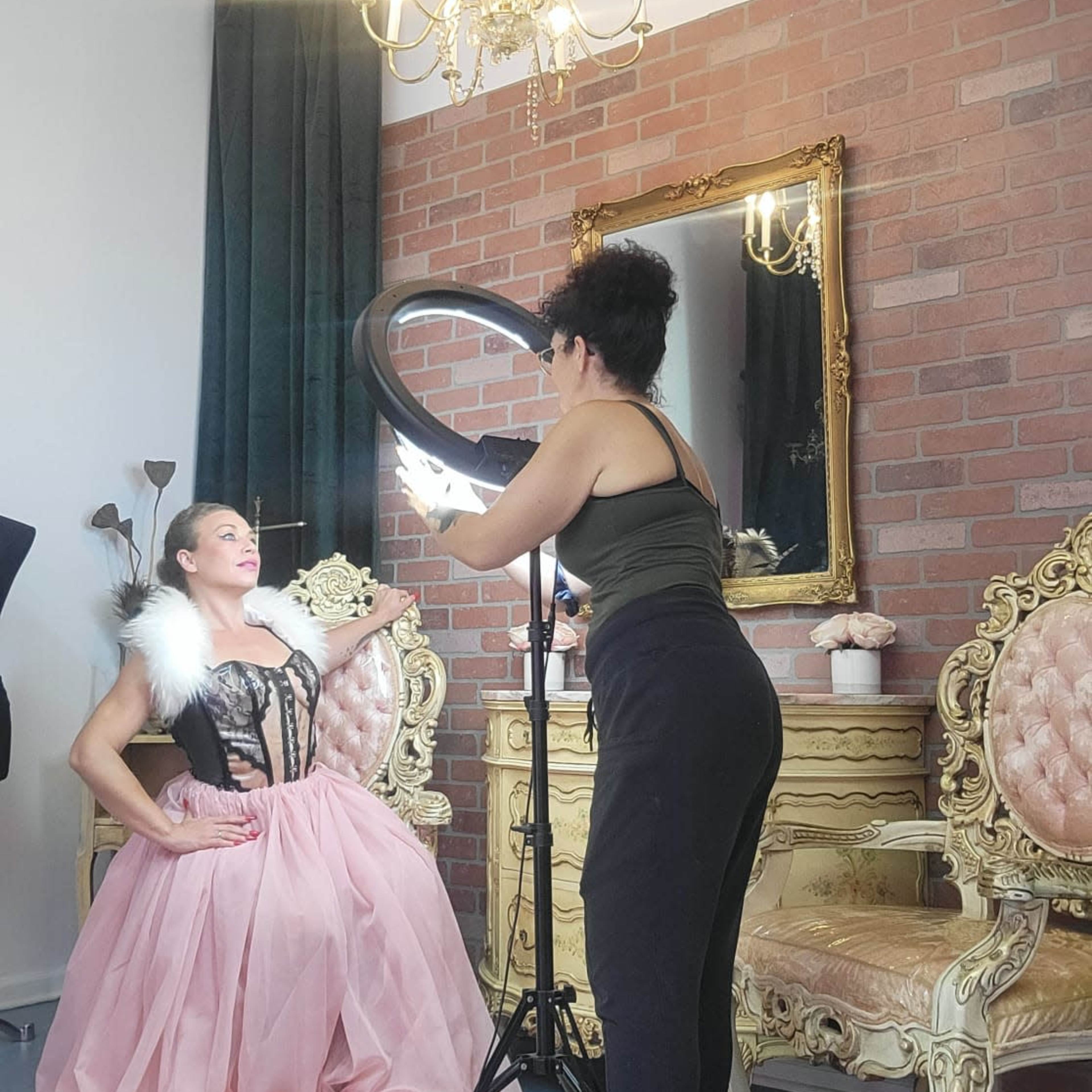 A woman in a pink gown poses while a stylist adjusts lighting equipment in a warmly decorated photo studio.