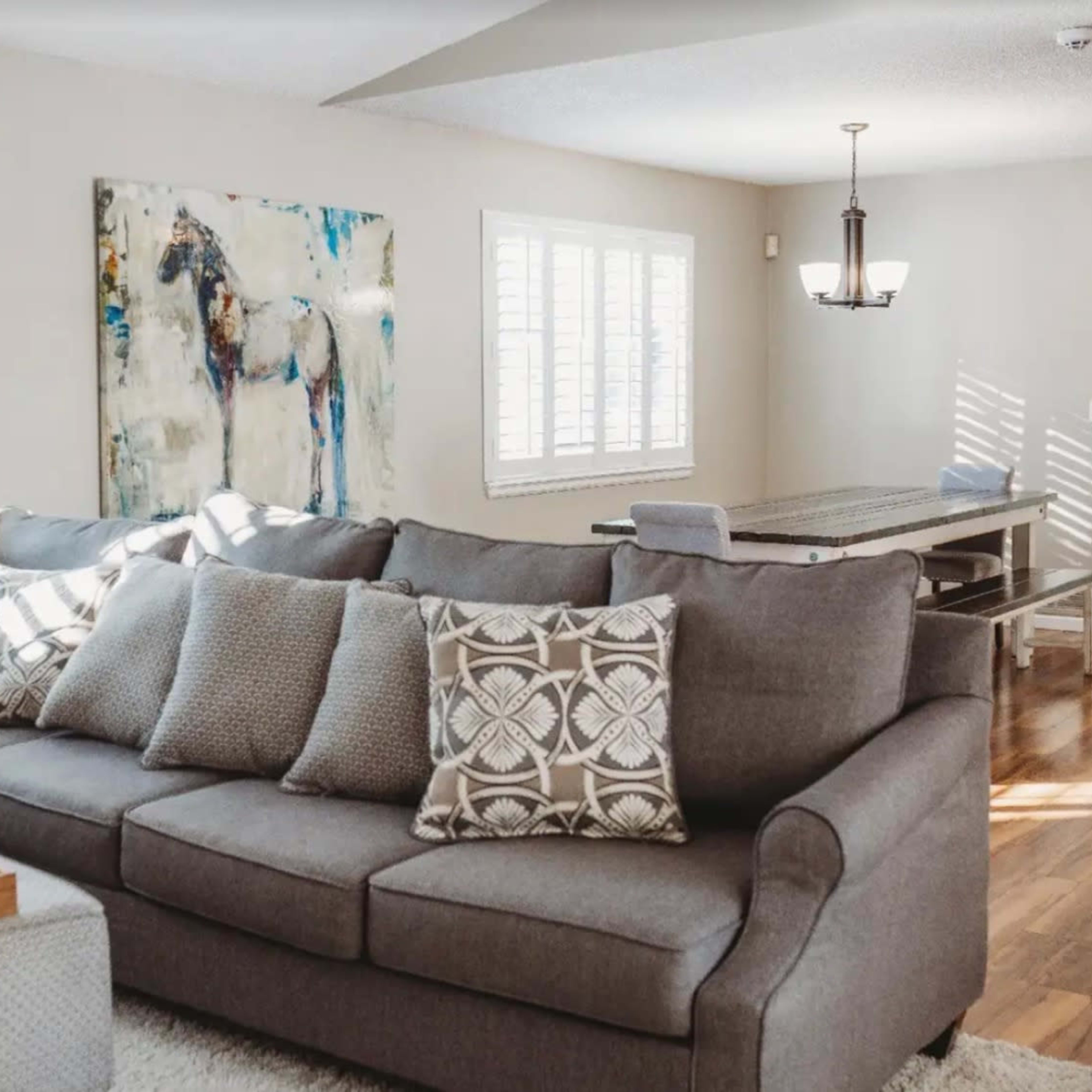 A living room features a gray sectional sofa with patterned pillows, an artwork on the wall, and a dining table in the background.