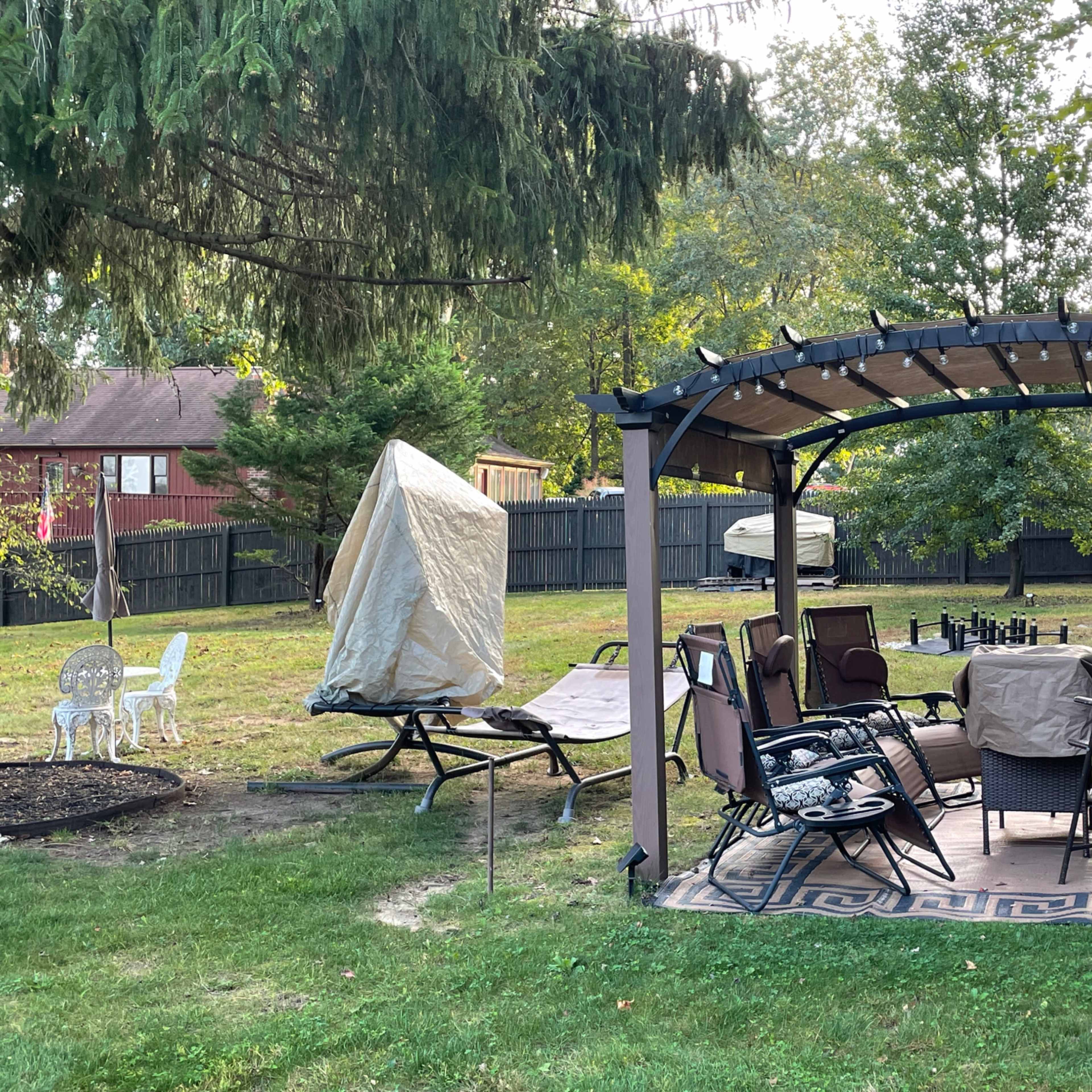 A backyard patio features a covered seating area with a table and chairs surrounded by trees and a grassy lawn.