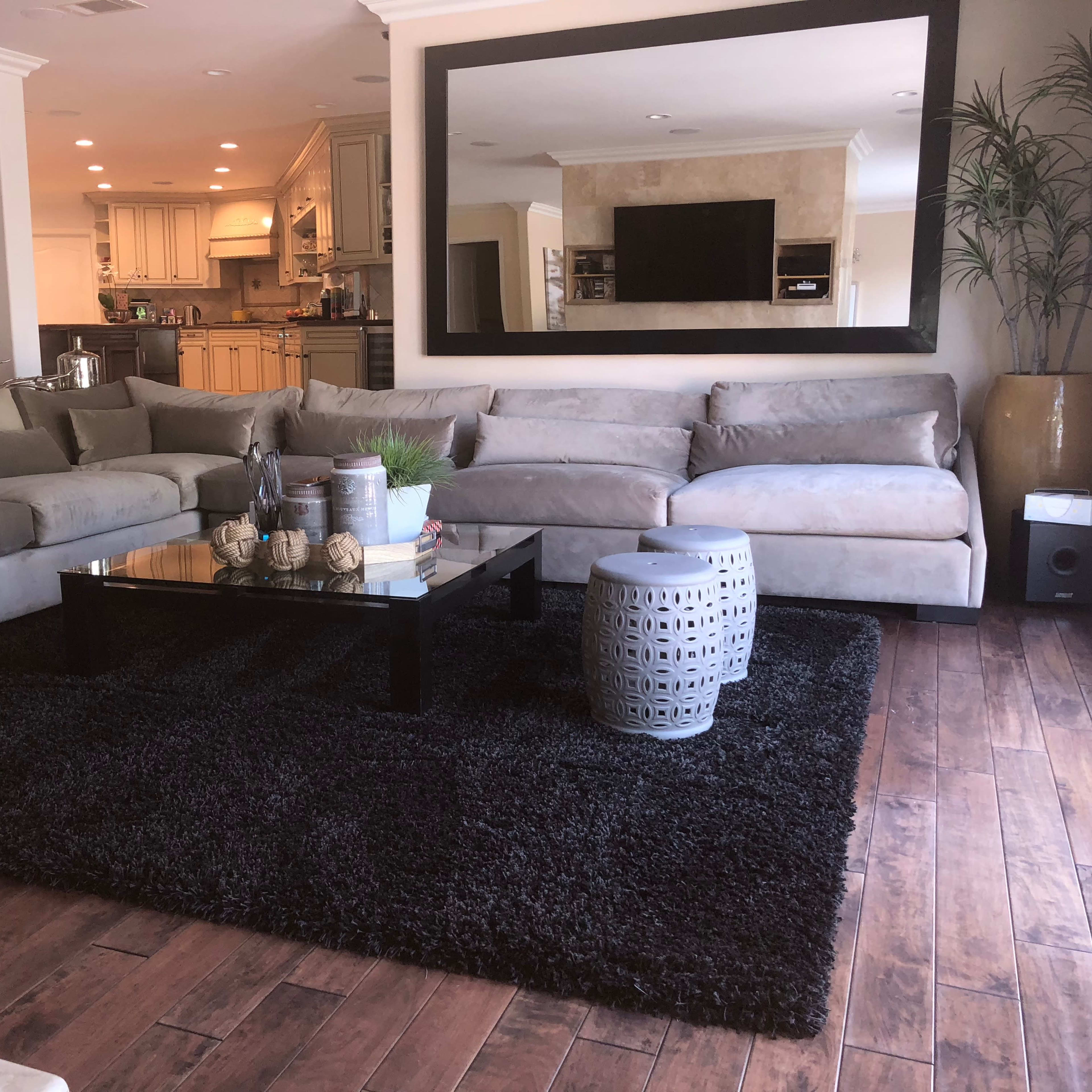 A modern living room featuring a large gray sectional sofa, a black coffee table on a textured rug, and an open kitchen in the background.