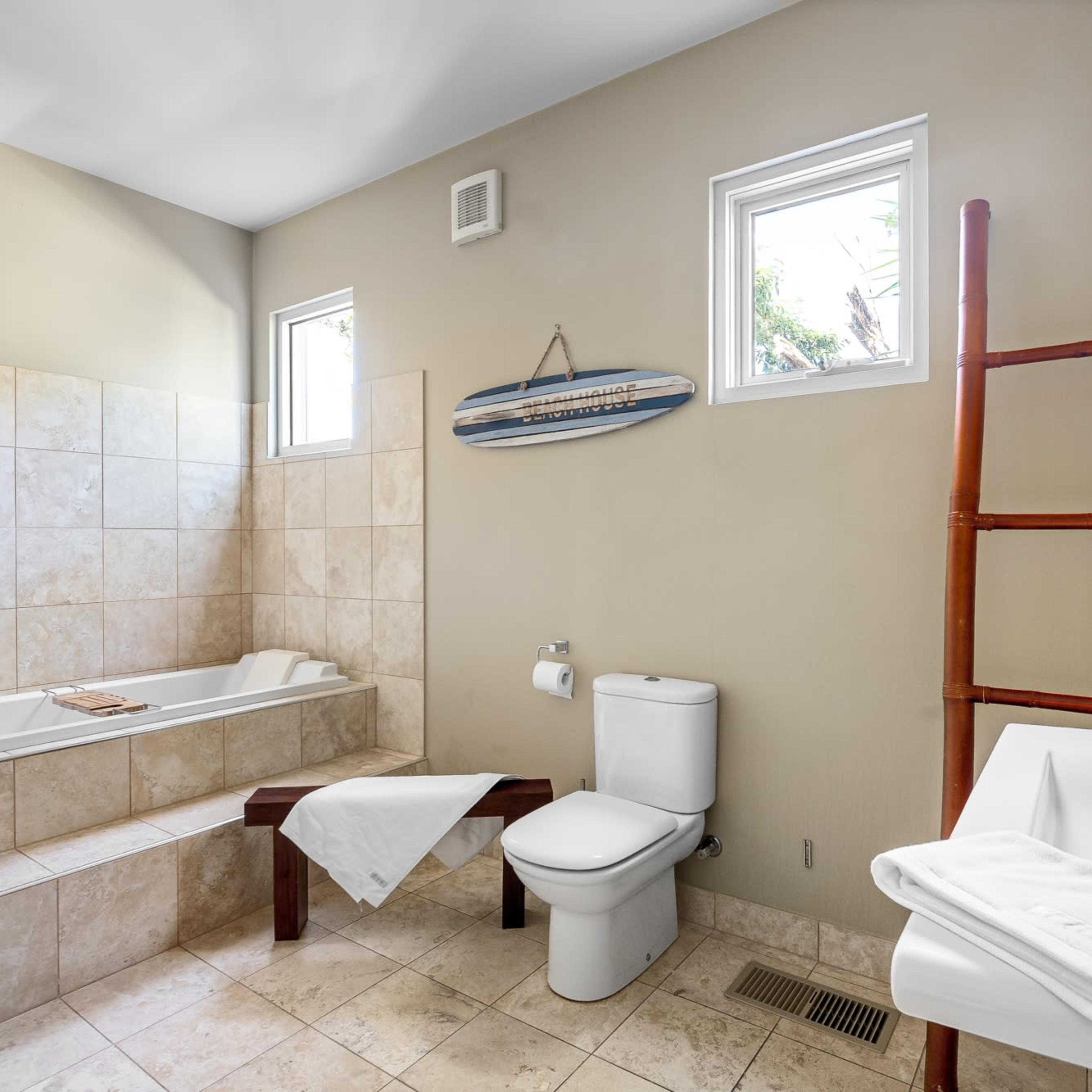 A light-colored bathroom featuring a bathtub, a white toilet, a sink with a mirror, and a wooden ladder for storage.