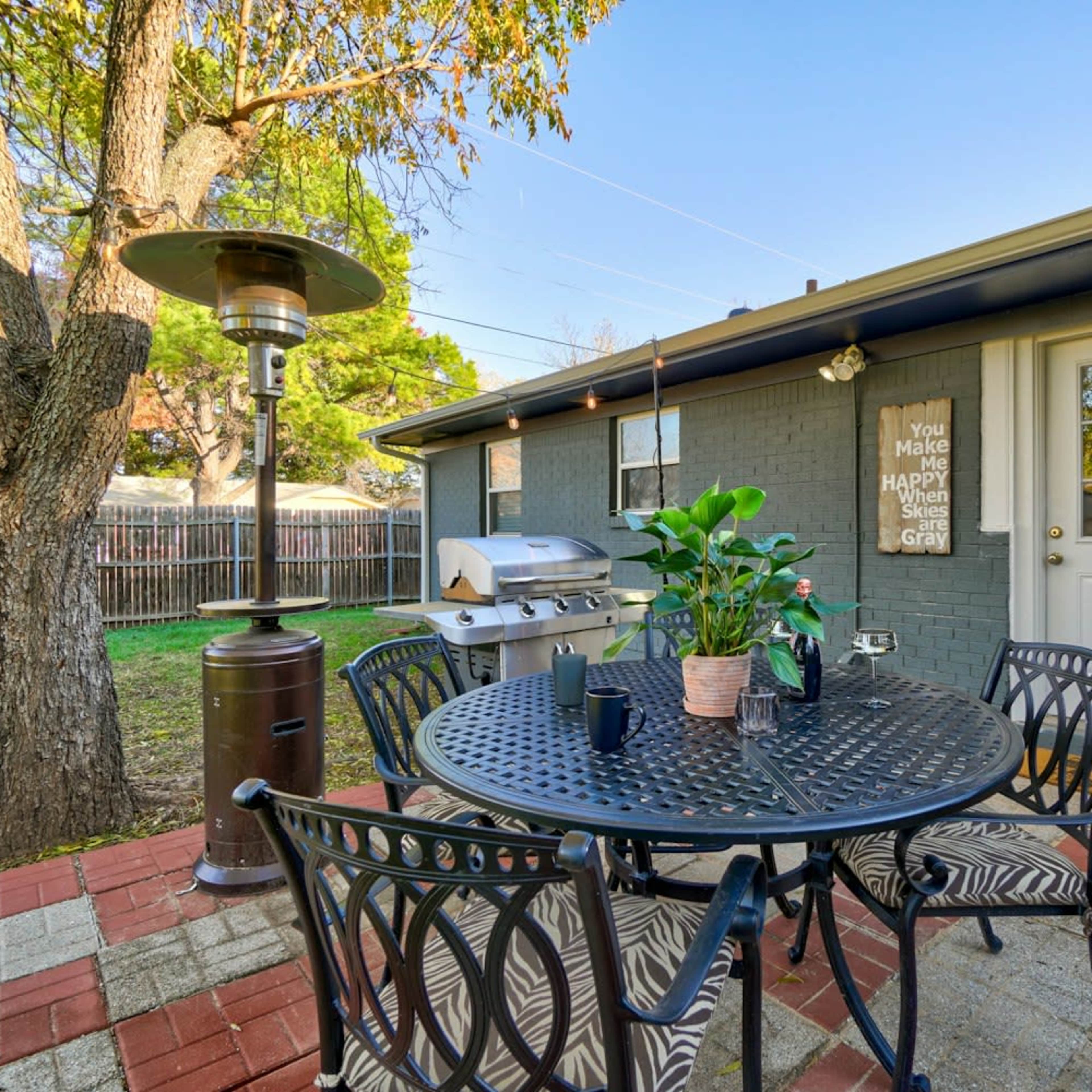 A patio with a round table, chairs, and a grill is set against a house with a door and string lights, surrounded by greenery.