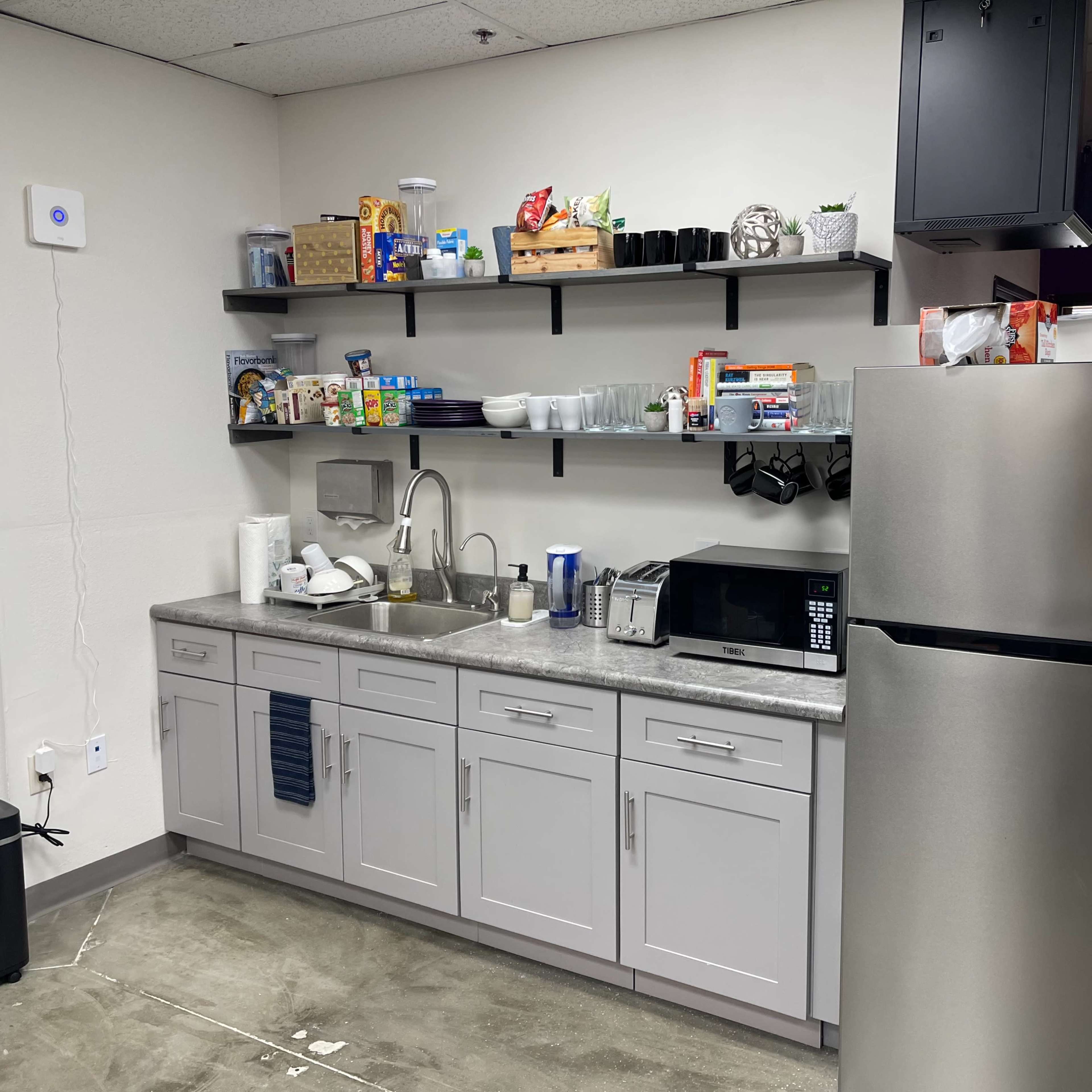A kitchen area with gray cabinets, a stainless steel refrigerator, a microwave, and open shelves stocked with various food items and dishware.