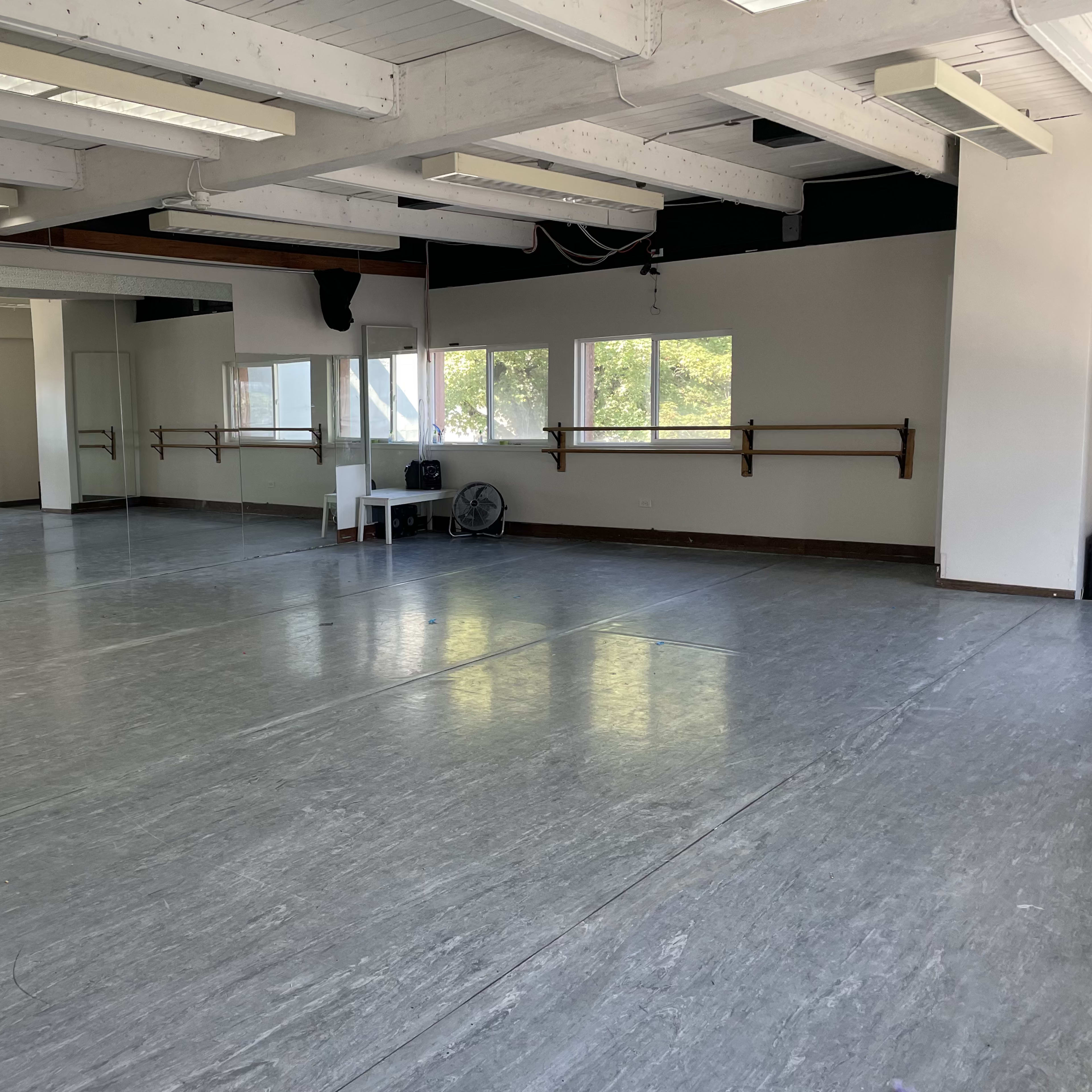 A spacious empty dance studio with wooden ballet barres, large windows, and a reflective gray floor.