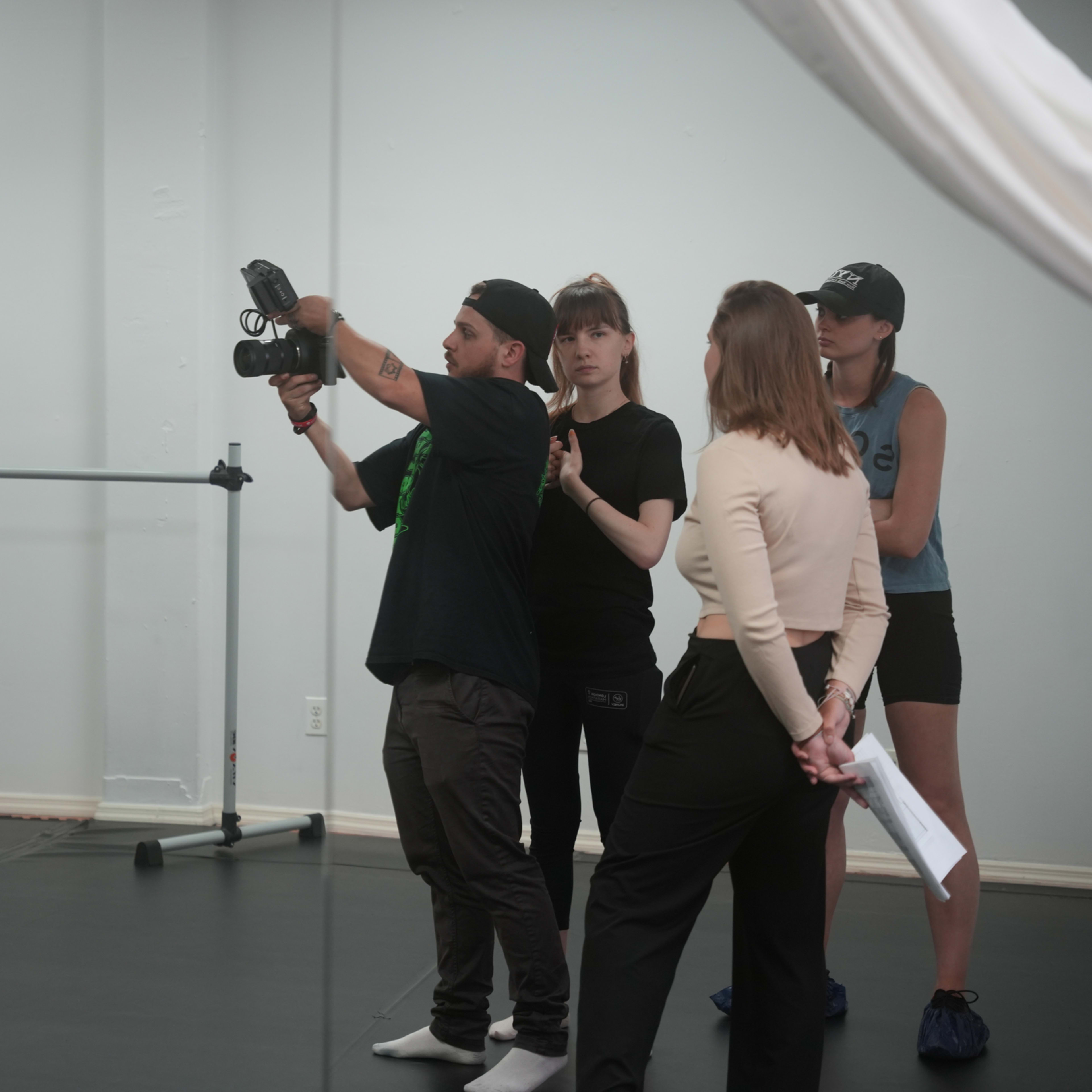 A young woman in a black crop top and leggings poses in front of a mirror while a group of three people, including a man with a camera, observes her in a dance studio.