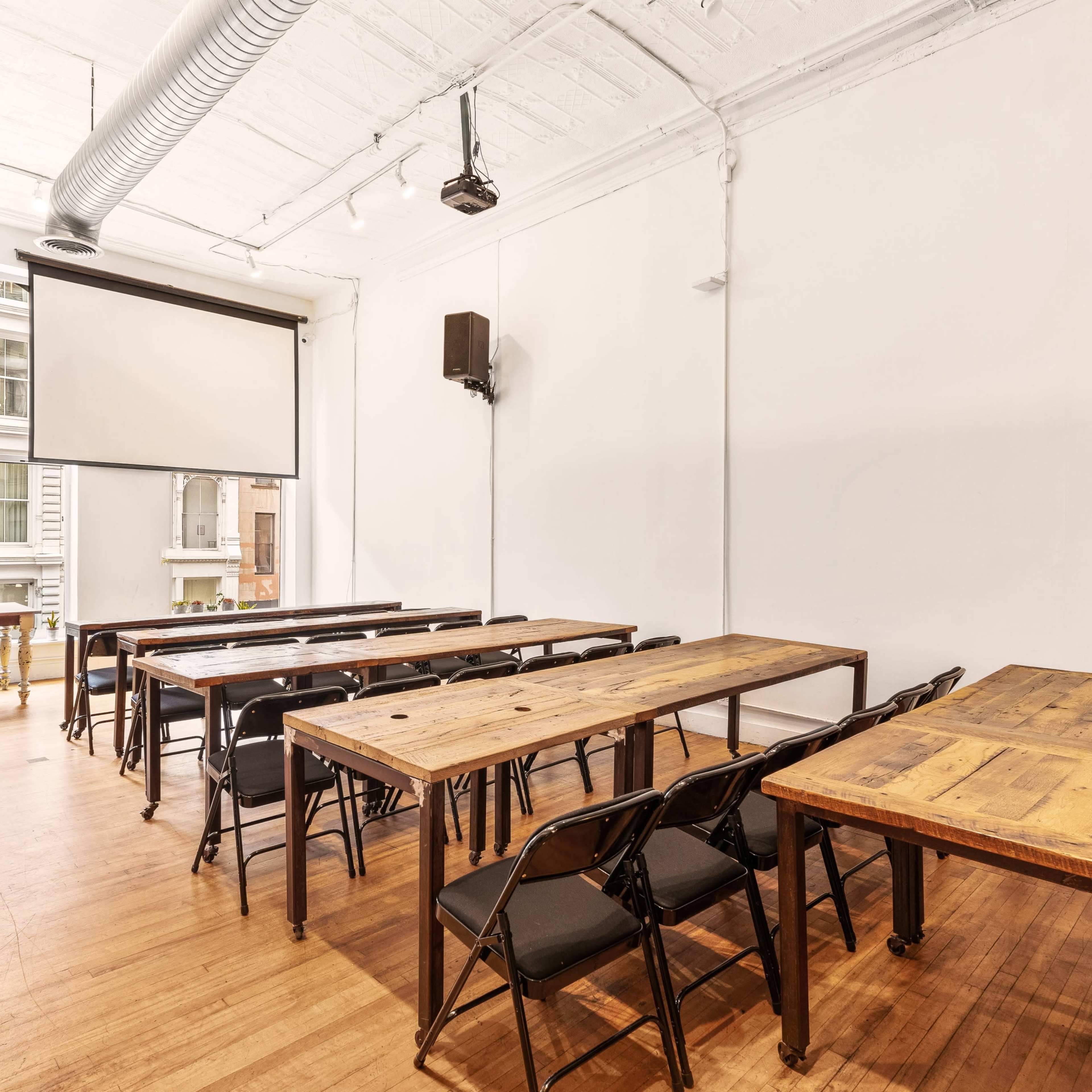 A spacious room with wooden tables arranged for a meeting, featuring a projector screen on the wall and large windows allowing natural light.