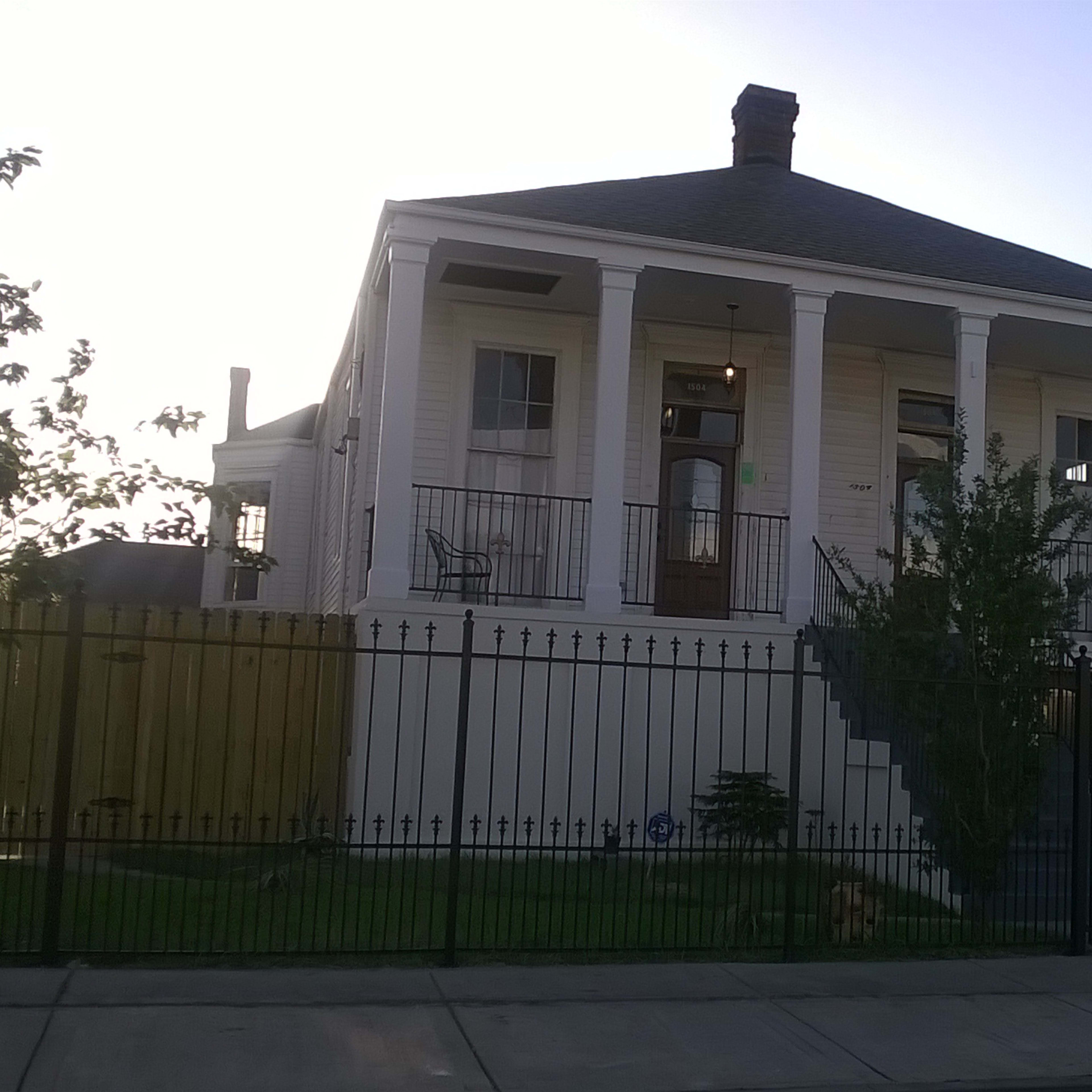 A two-story white house with a front porch supported by four columns, surrounded by a black wrought-iron fence.