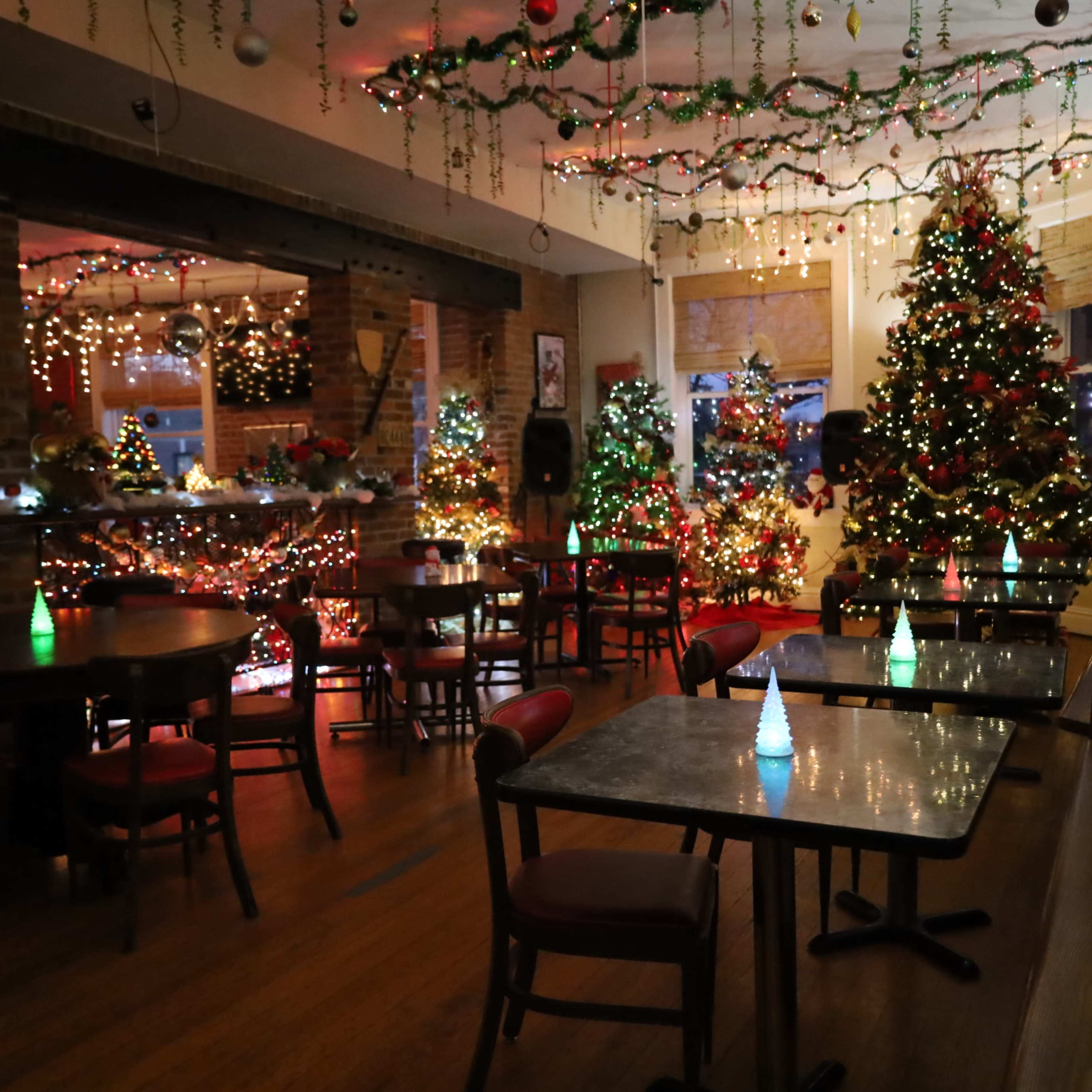 A warmly decorated indoor space with several Christmas trees adorned with lights and festive decorations, surrounded by dining tables and chairs.