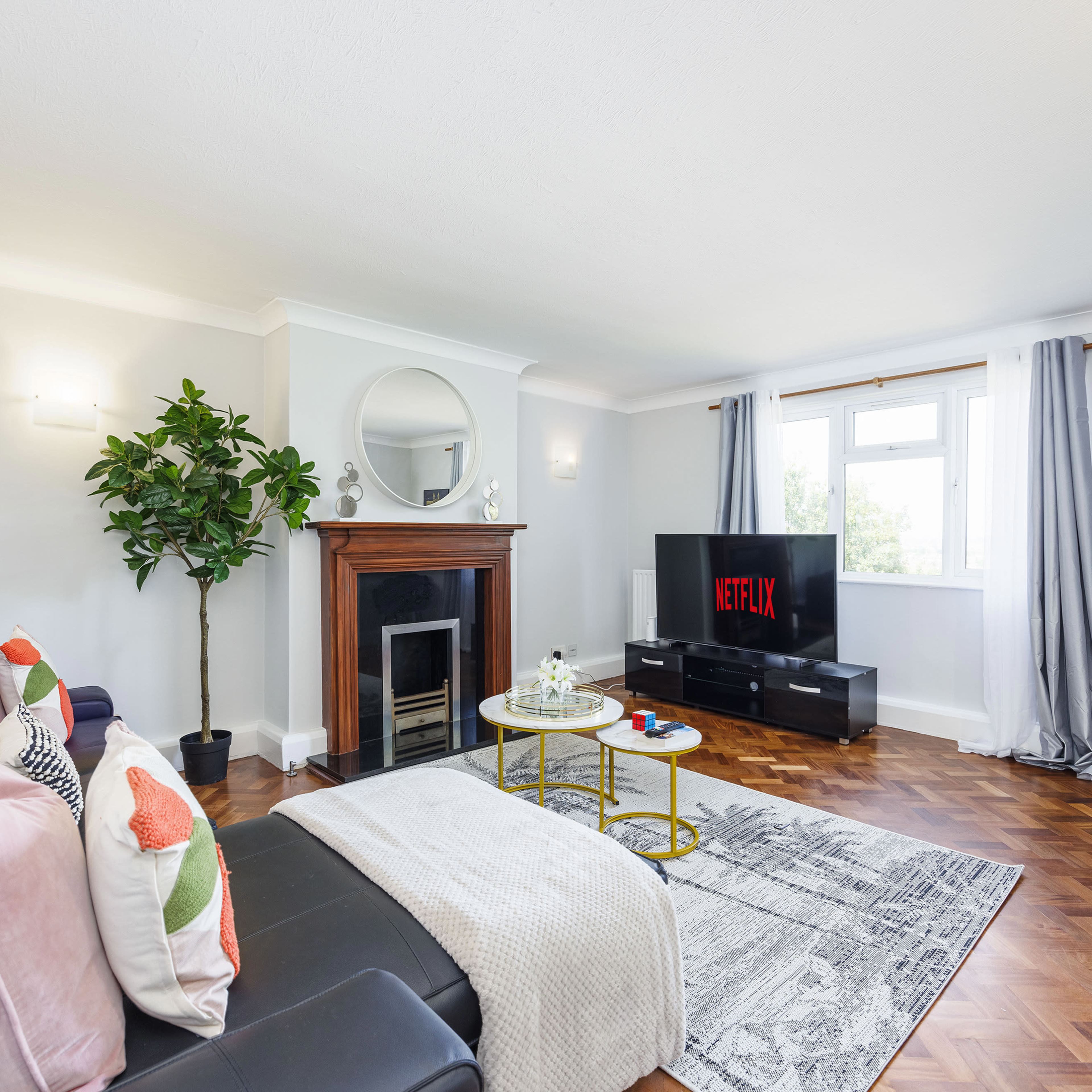 The living room features a black leather sofa with decorative pillows, a television displaying the Netflix logo, a round mirror above a fireplace, and a potted plant.