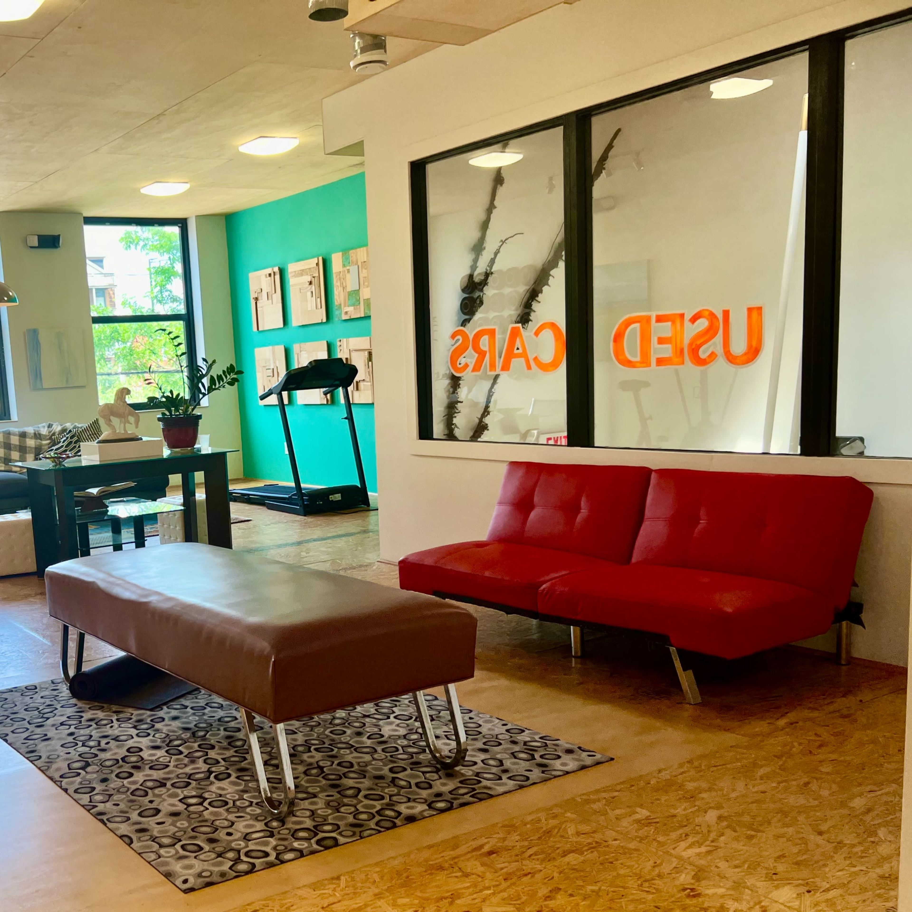 A spacious, modern workspace with a red couch, a wooden table, exercise equipment, and large windows displaying the words "USED CARS" in bright orange.