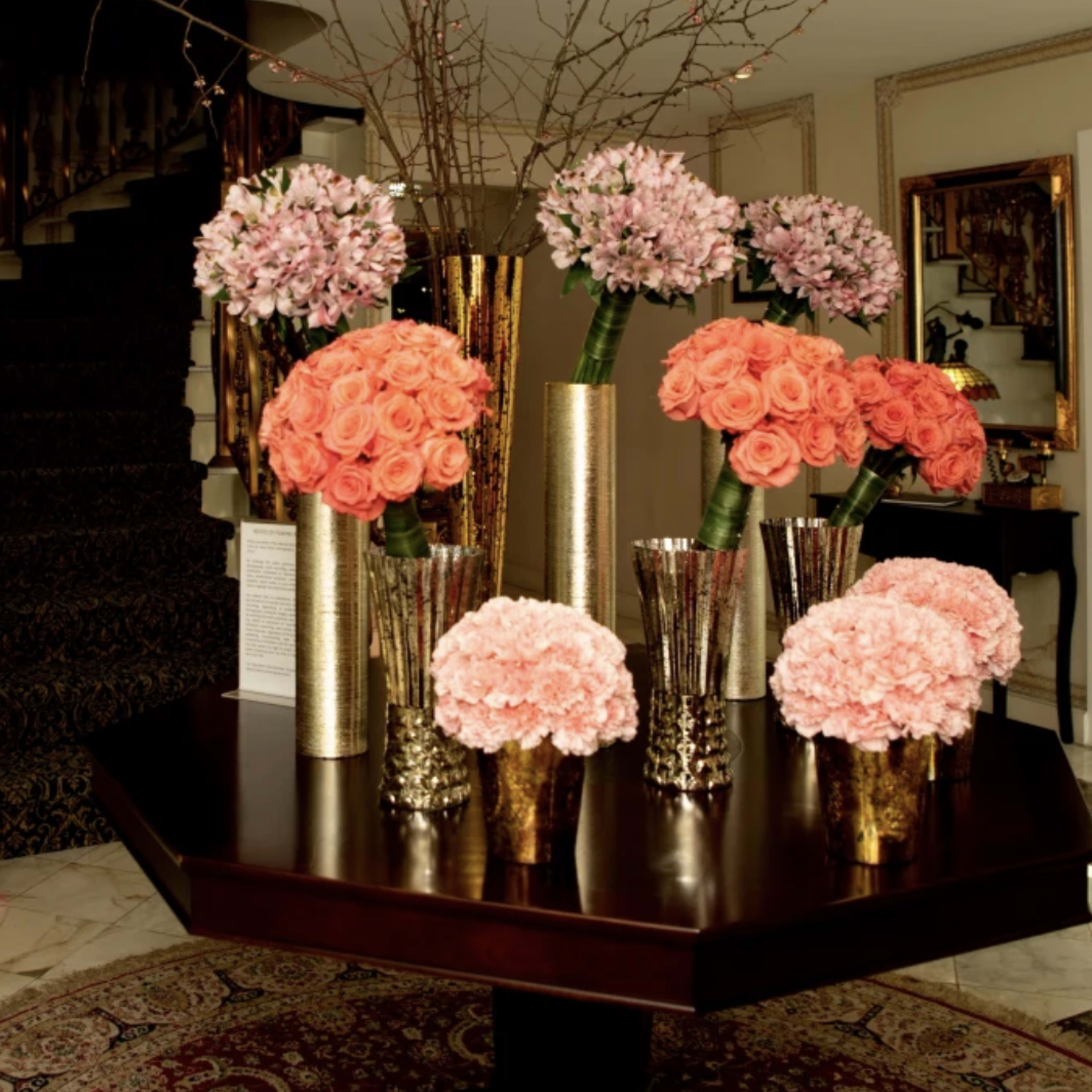 A foyer features a round wooden table with vibrant flower arrangements in metallic vases and a staircase in the background.