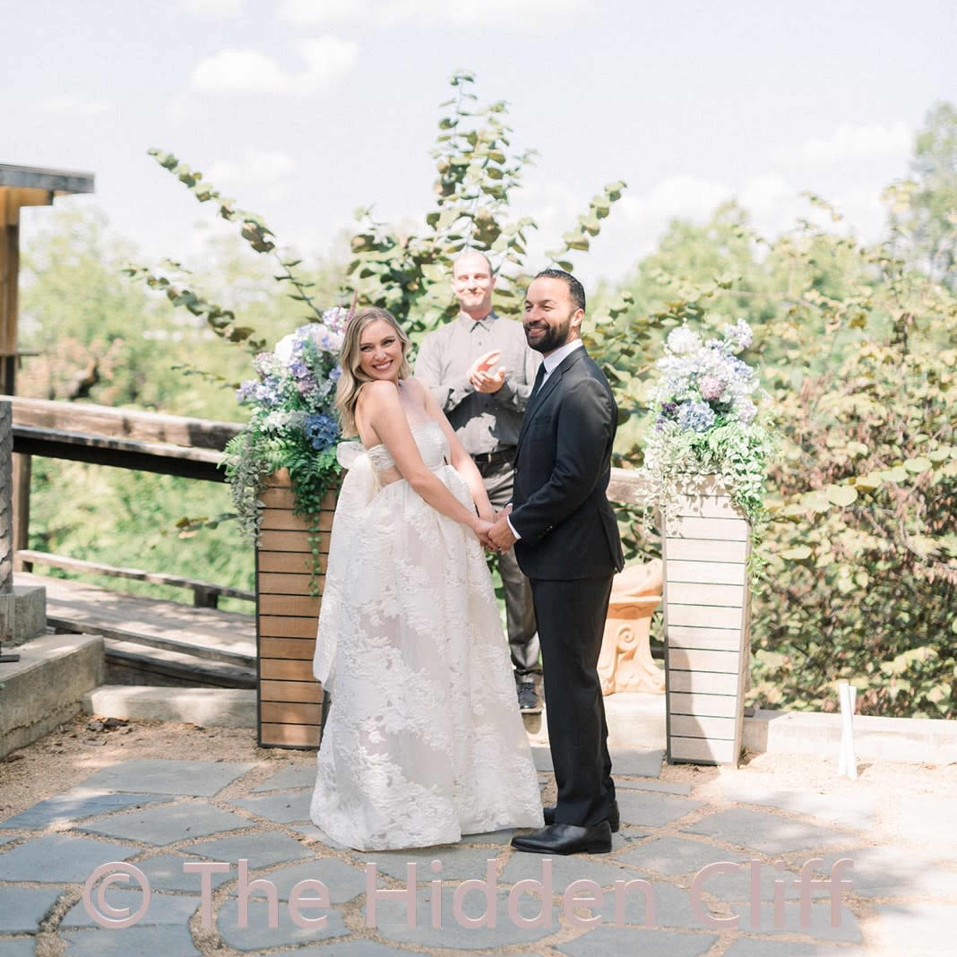A bride and groom stand together during their wedding ceremony, with a celebrant in the background amidst floral arrangements and greenery.