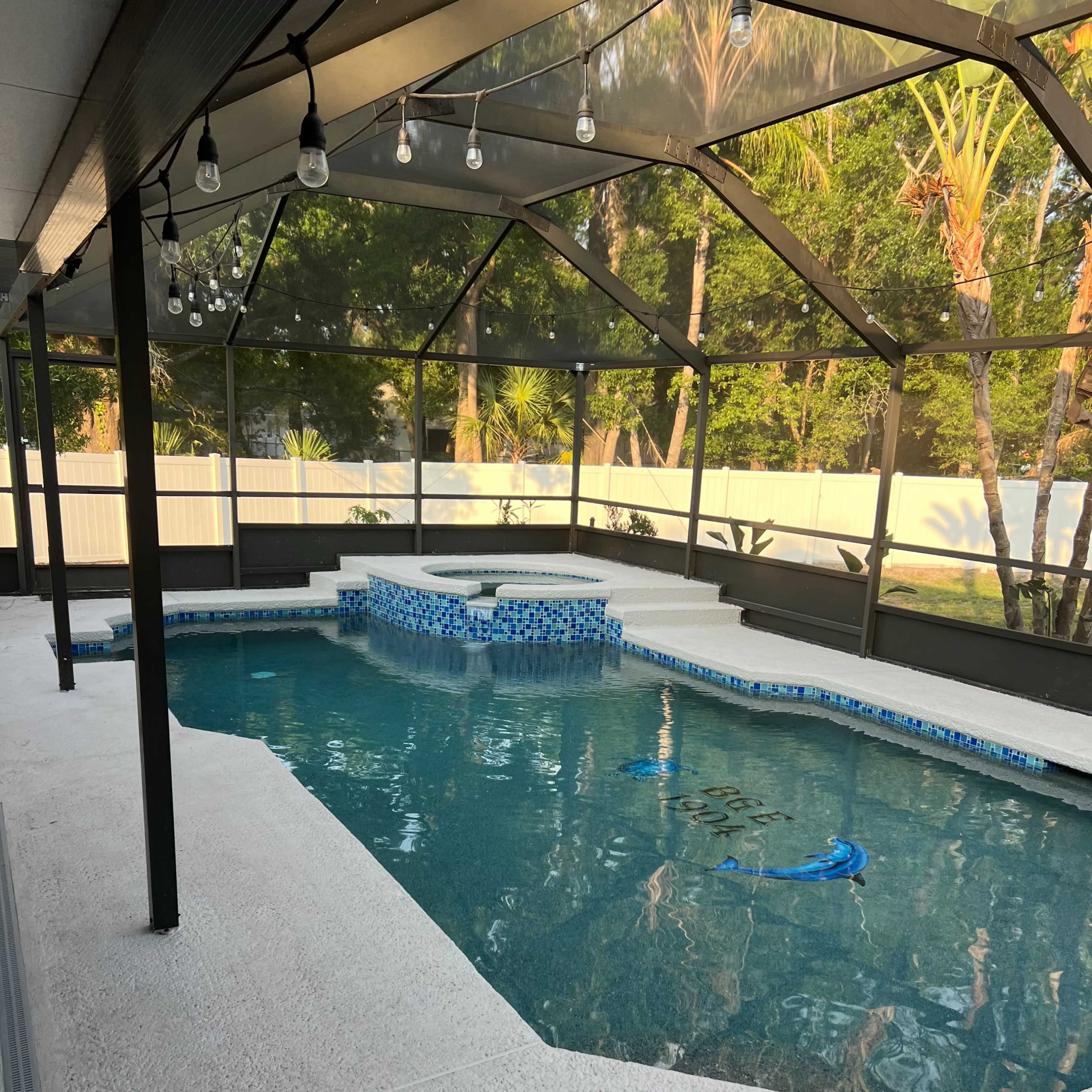 A screened-in swimming pool area featuring a blue-tiled spa and white deck surrounded by lush greenery.