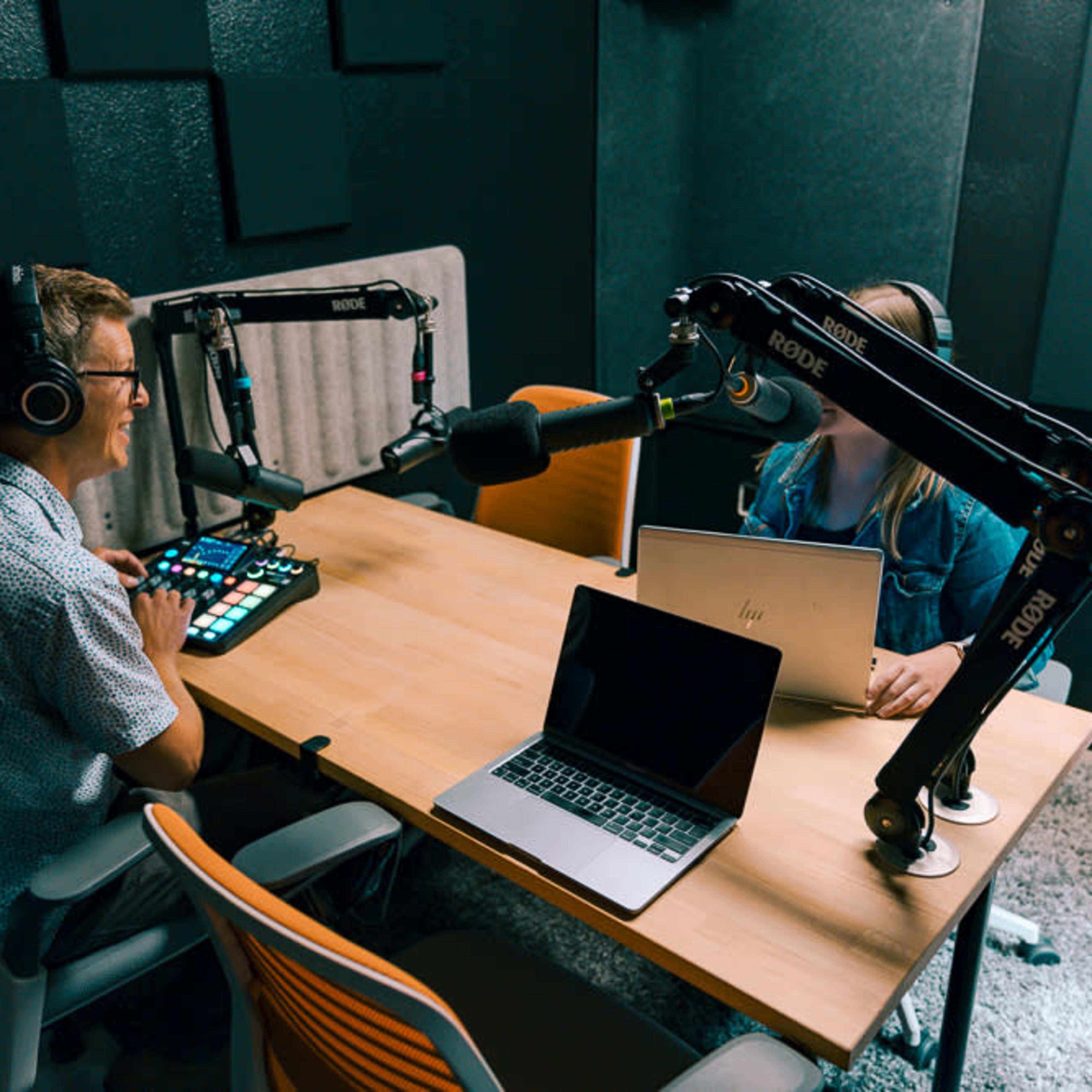 Two people sit at a table in a recording studio, equipped with microphones and laptops, preparing for a podcast session.