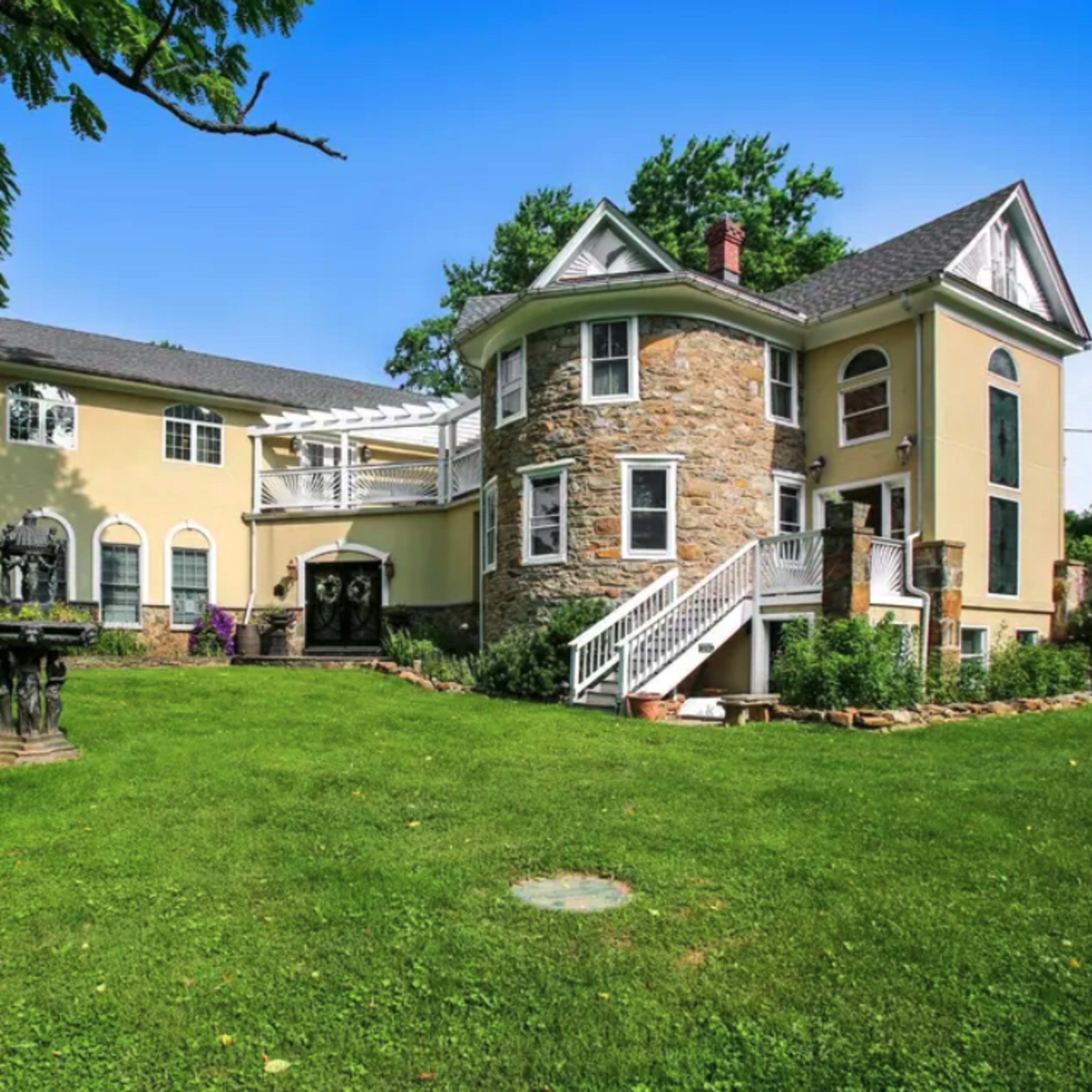 A two-story house with a circular stone tower, a large deck, and a well-maintained lawn surrounded by trees.