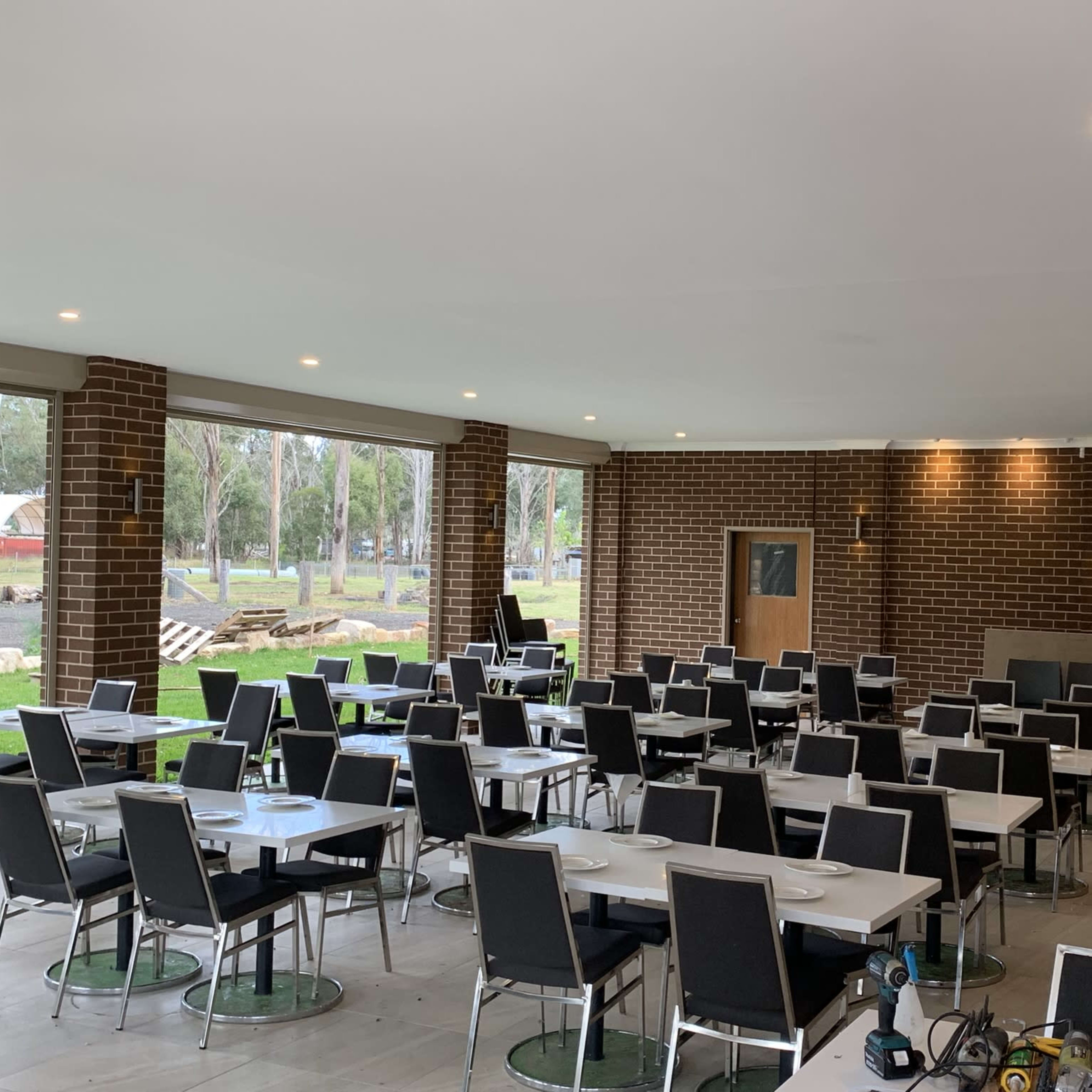 A spacious dining area with multiple tables and chairs arranged systematically, and large windows providing a view of the outside greenery.