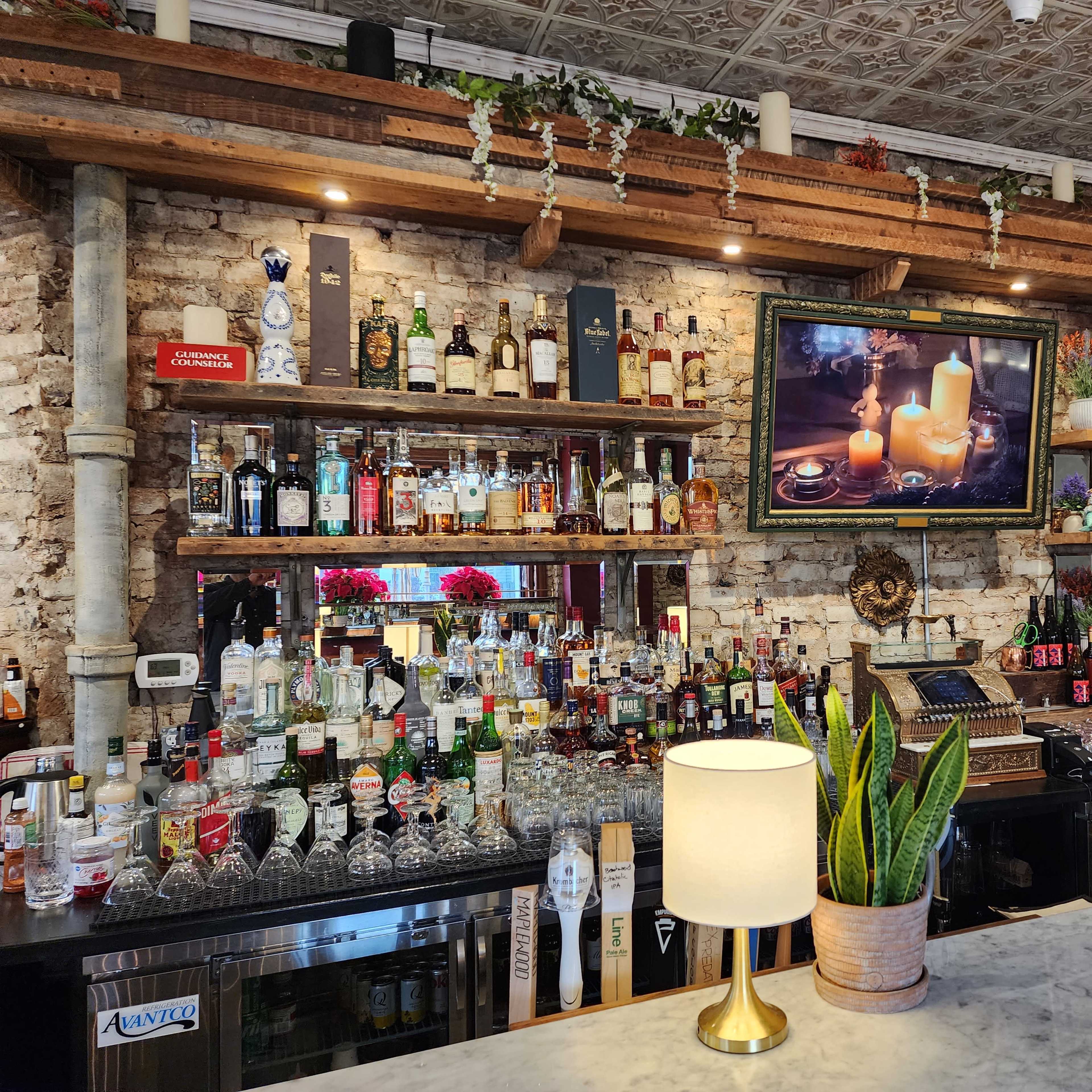 A well-stocked bar with a wooden countertop, shelves filled with various liquor bottles, and a television displaying a candlelit scene above the bar.