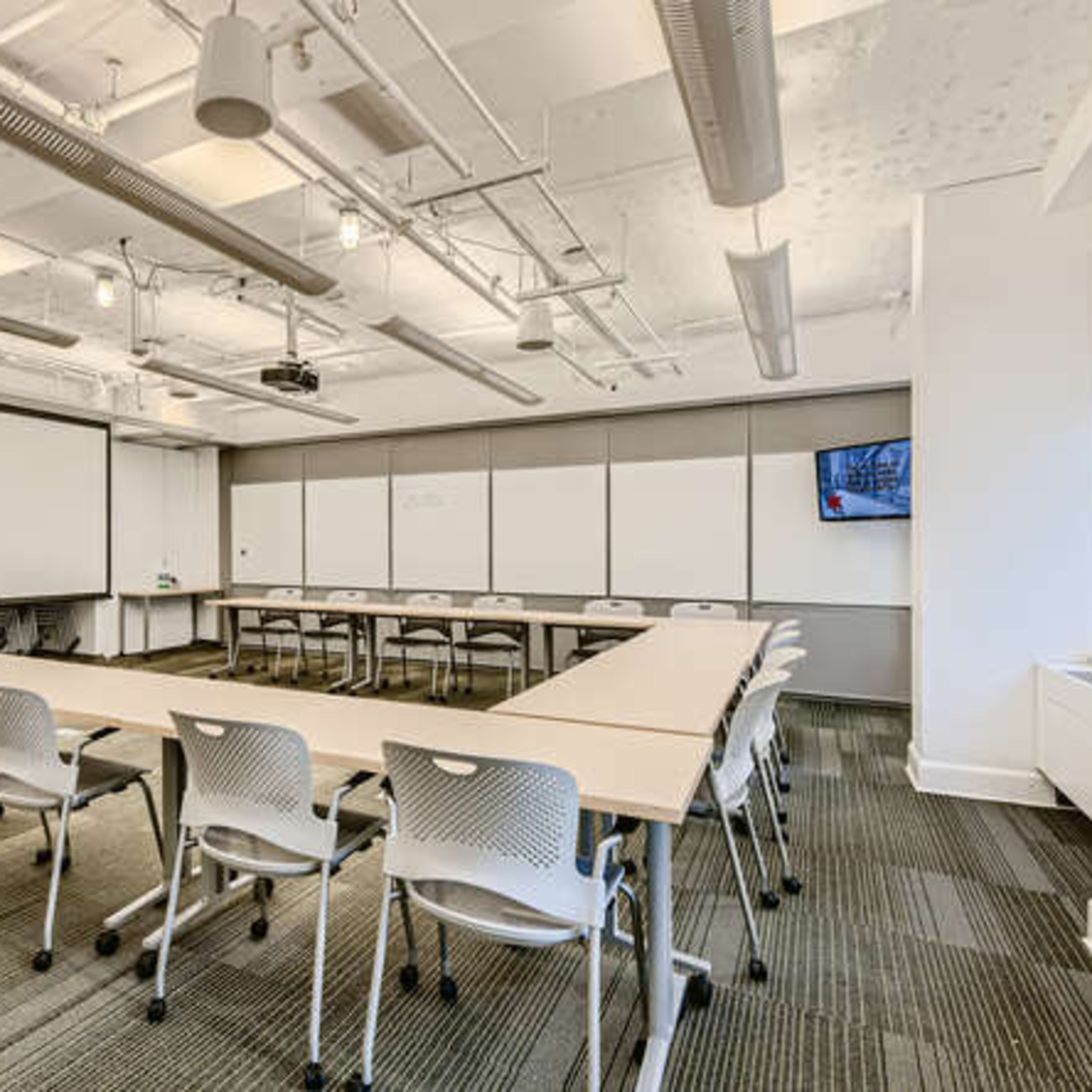 A conference room is arranged with a long table surrounded by chairs, featuring a projector screen and whiteboards along the walls.