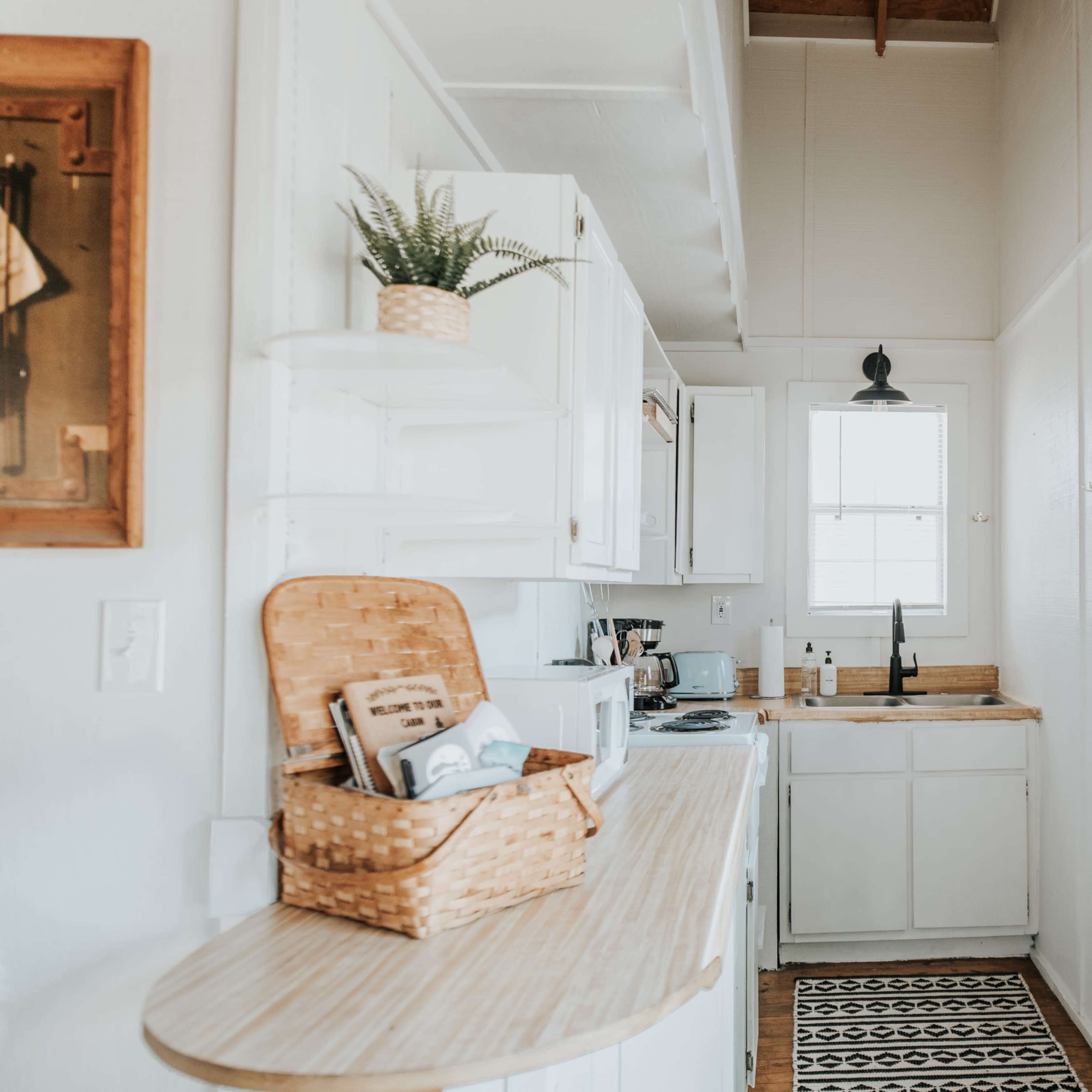A cozy kitchen features white cabinets, a sink under a window, and minimal decor, including a wall clock and a framed motivational poster.