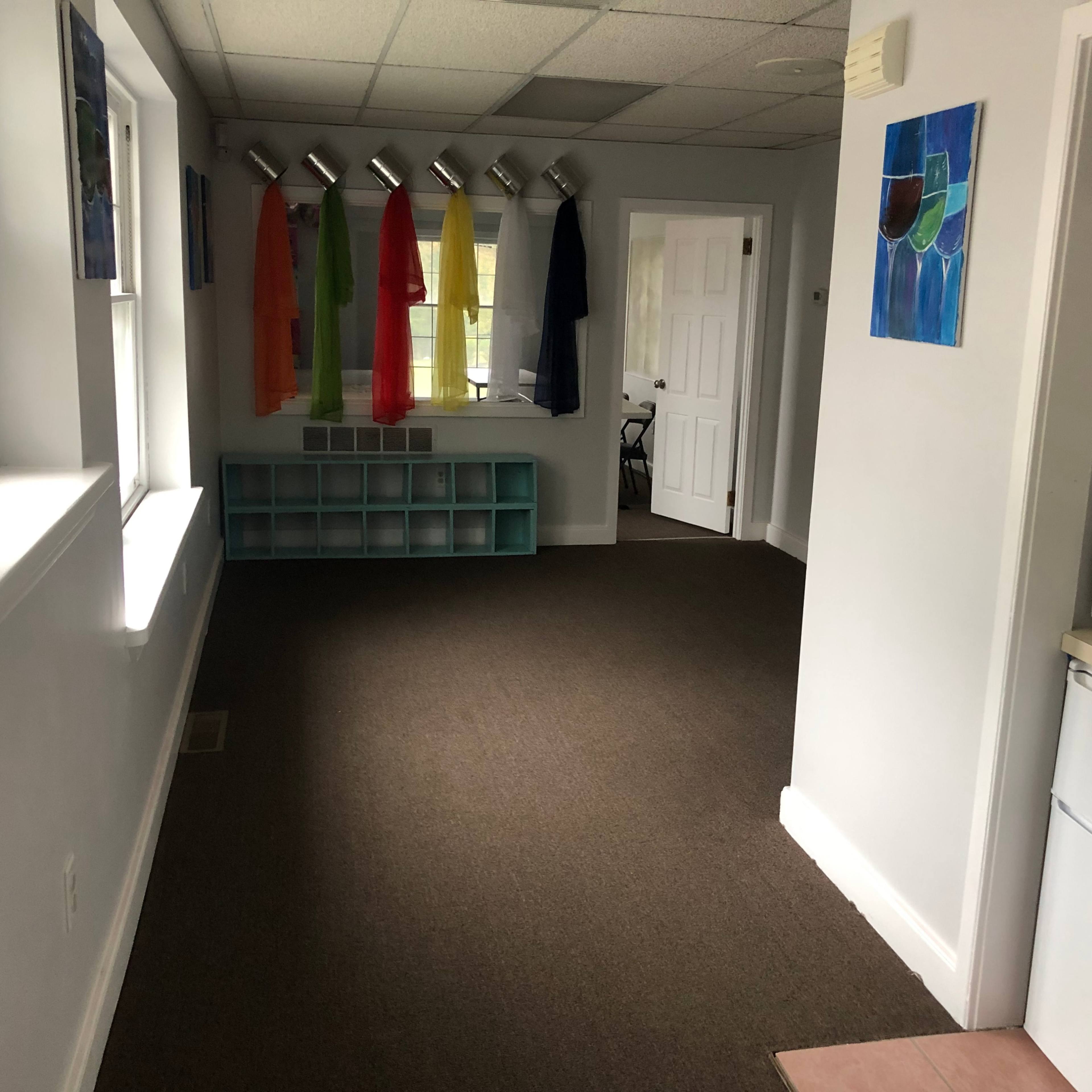 An interior room with a carpeted floor, white walls, a row of colored garments hanging from a rack, and a kitchenette area featuring a refrigerator and a microwave.