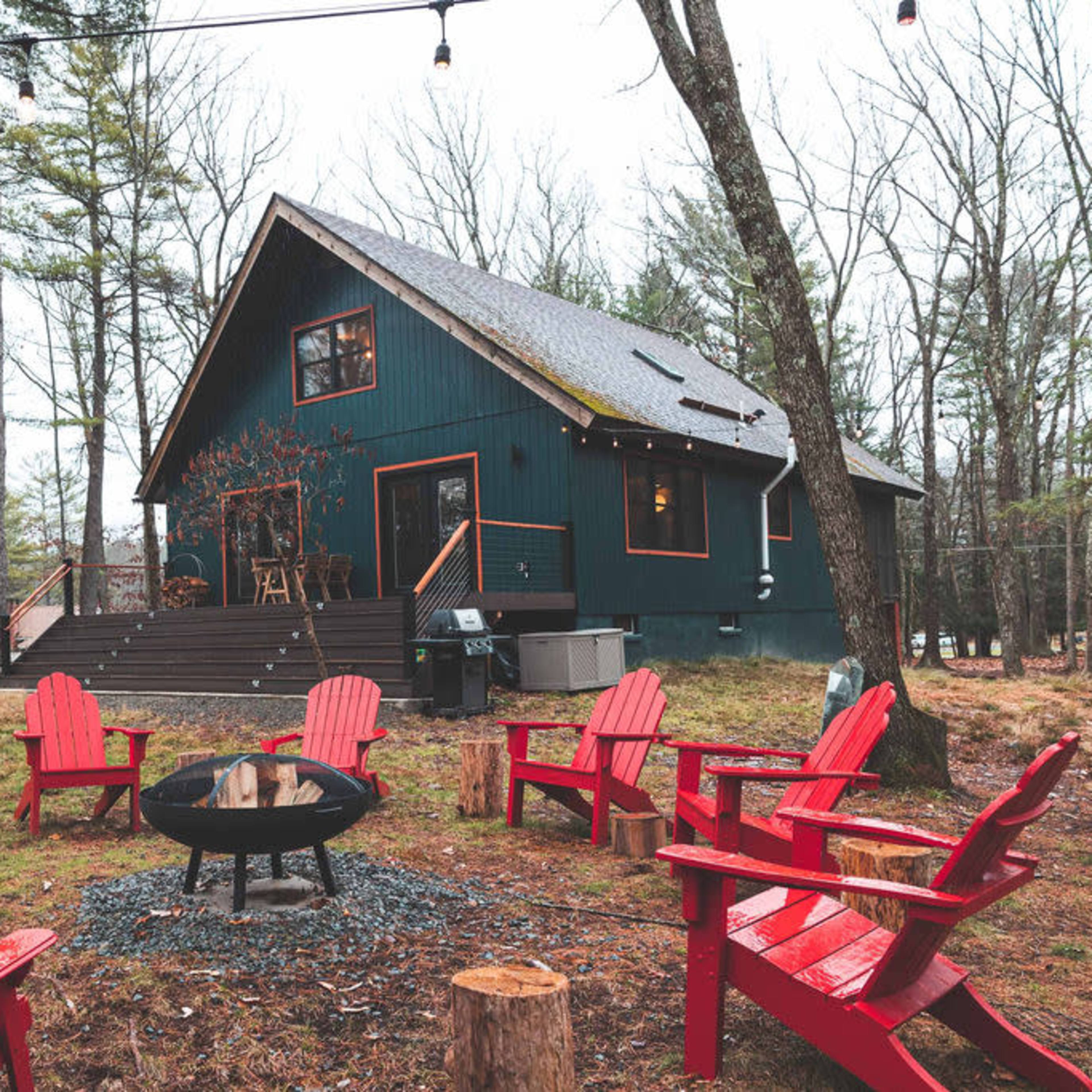 A green two-story cabin is surrounded by red Adirondack chairs and a fire pit in a wooded area.