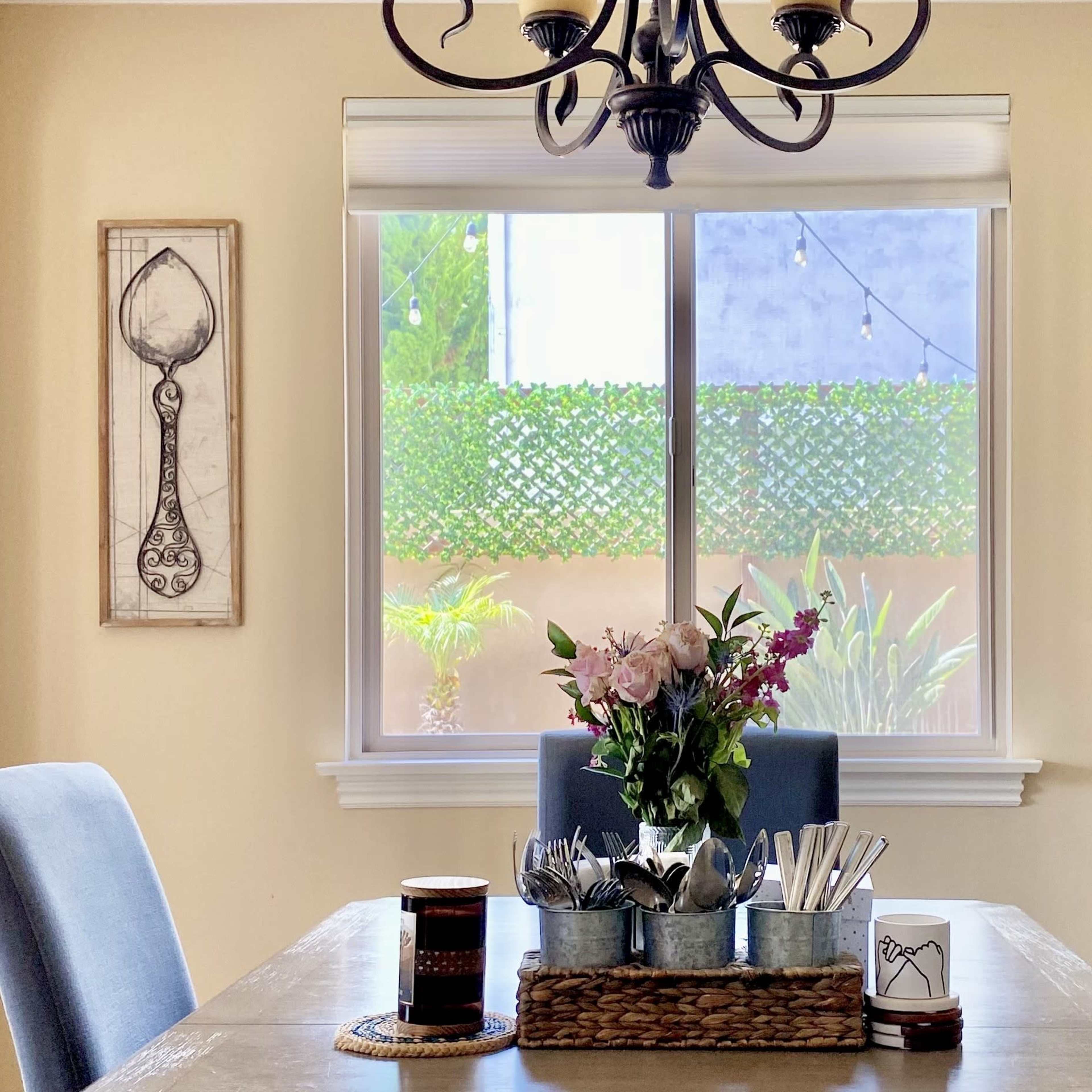 A dining table is set with a floral centerpiece and utensils on a tray, surrounded by chairs and framed drawings of a spoon and fork on the walls.