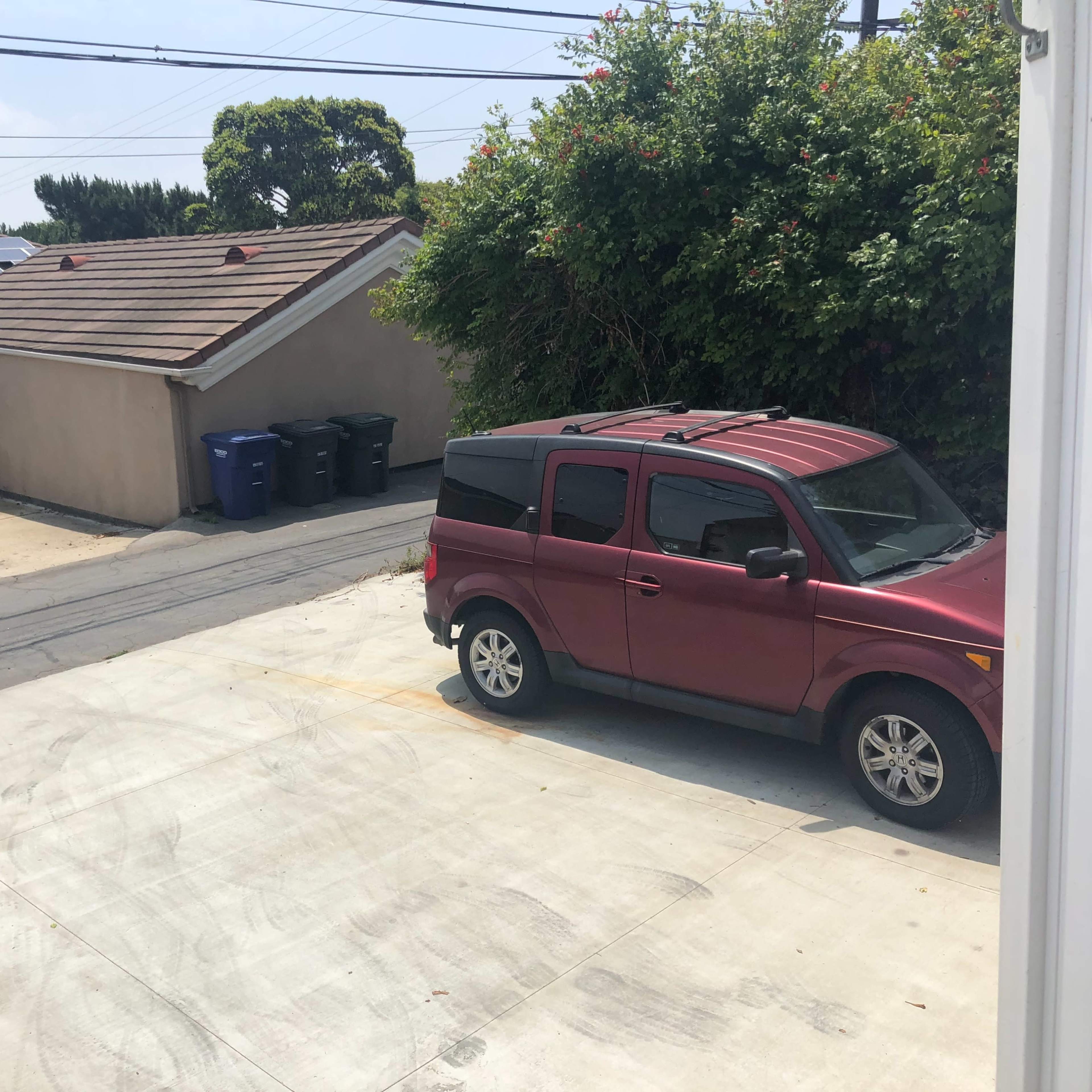 A burgundy vehicle is parked in a driveway alongside several recycling and trash bins, with a house and trees in the background.