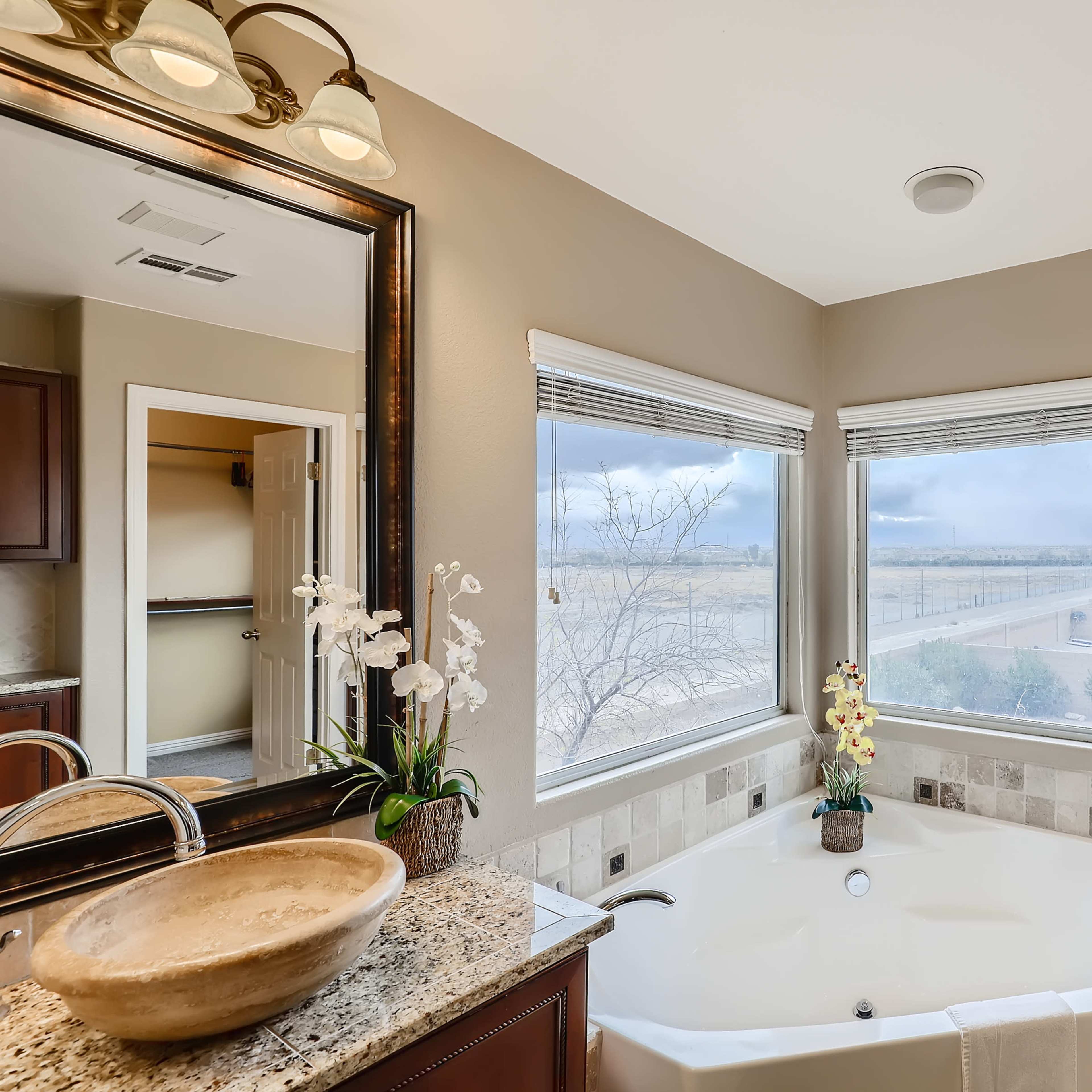 A bathroom with a large bathtub, double sinks, and two mirrors, featuring a window with a view of a cloudy landscape.
