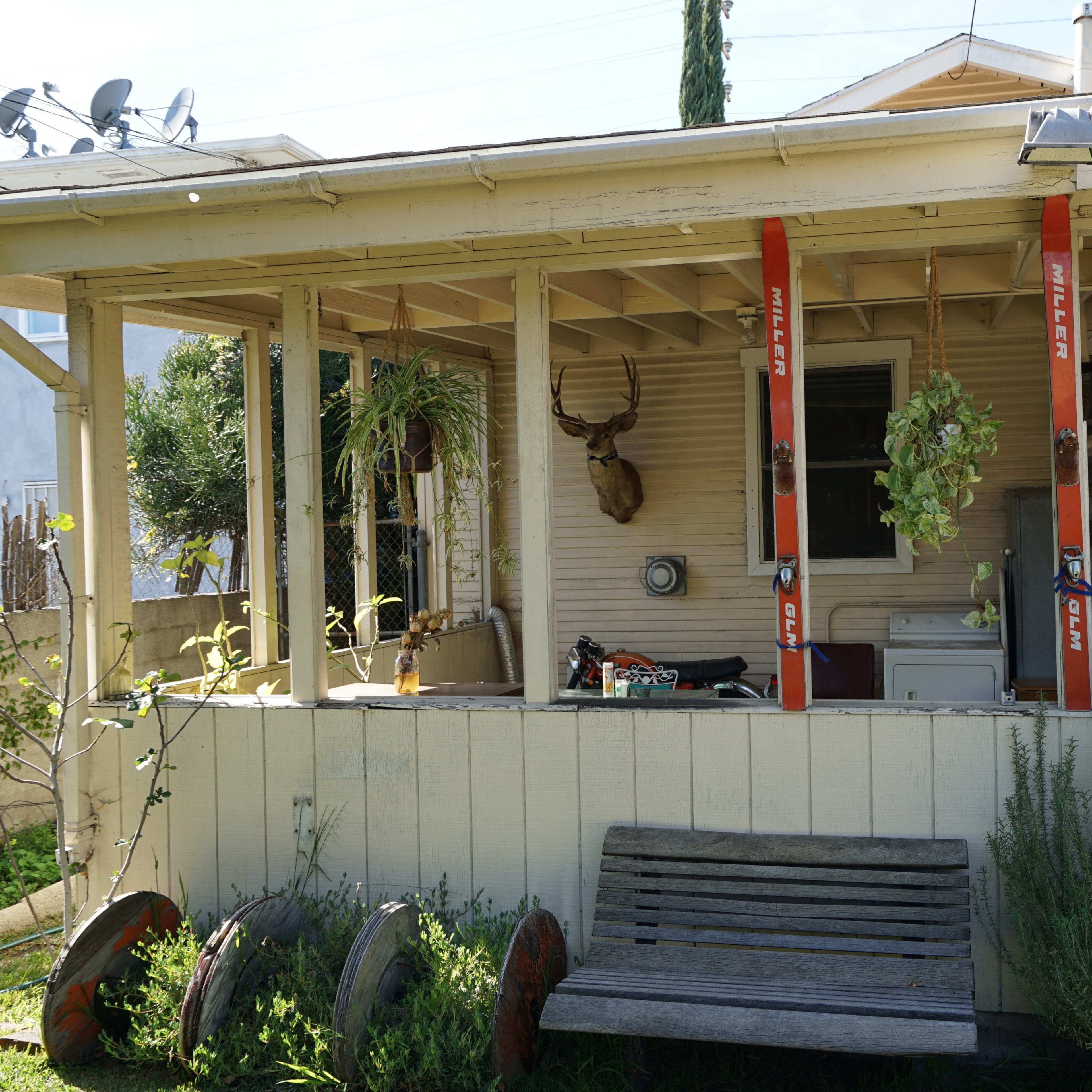 A porch with two vertical red posts, a mounted deer head, and a wooden bench is situated in front of a house surrounded by plants and equipment.