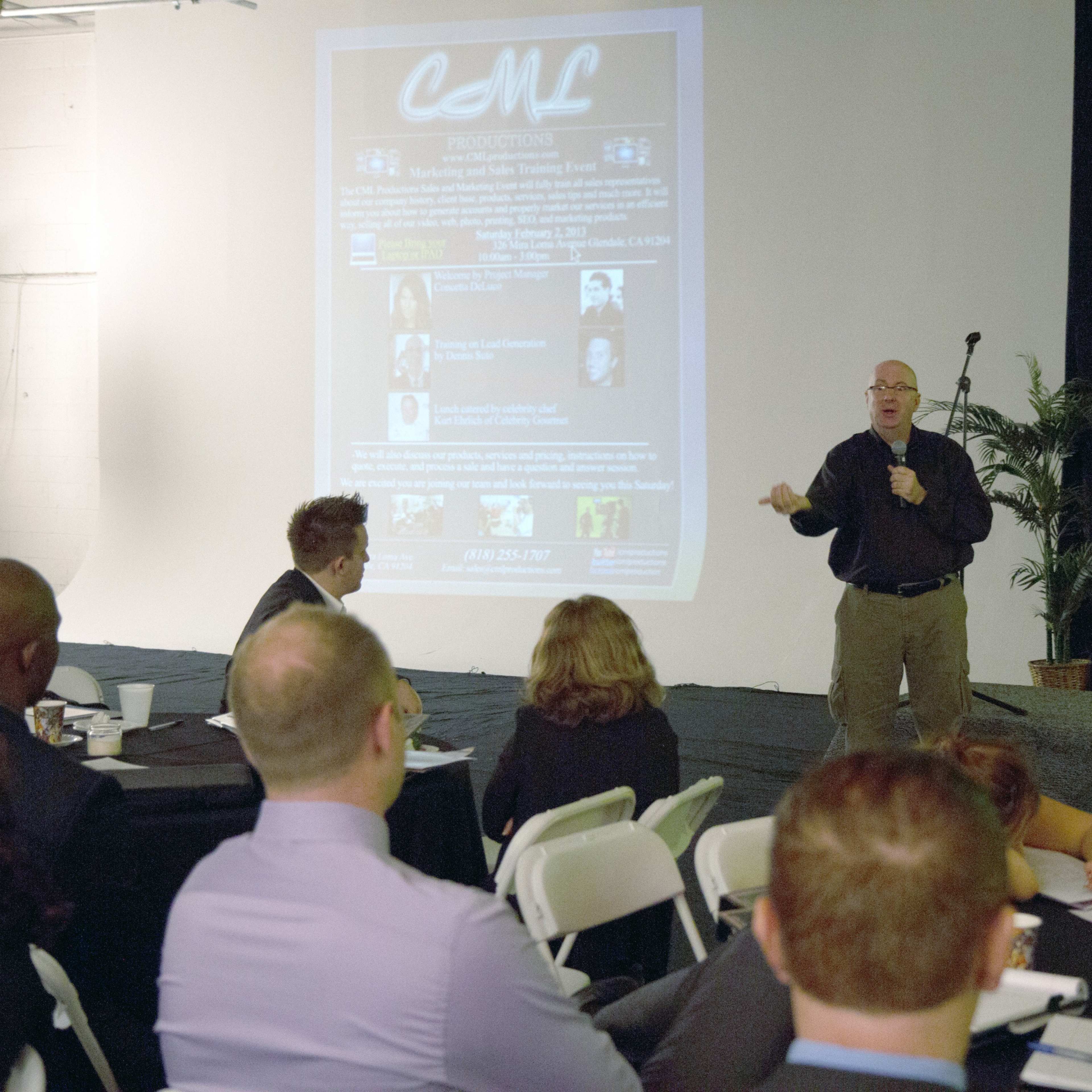 A speaker presents to an audience in a conference room, with a large projection screen displaying information about a production company in the background.