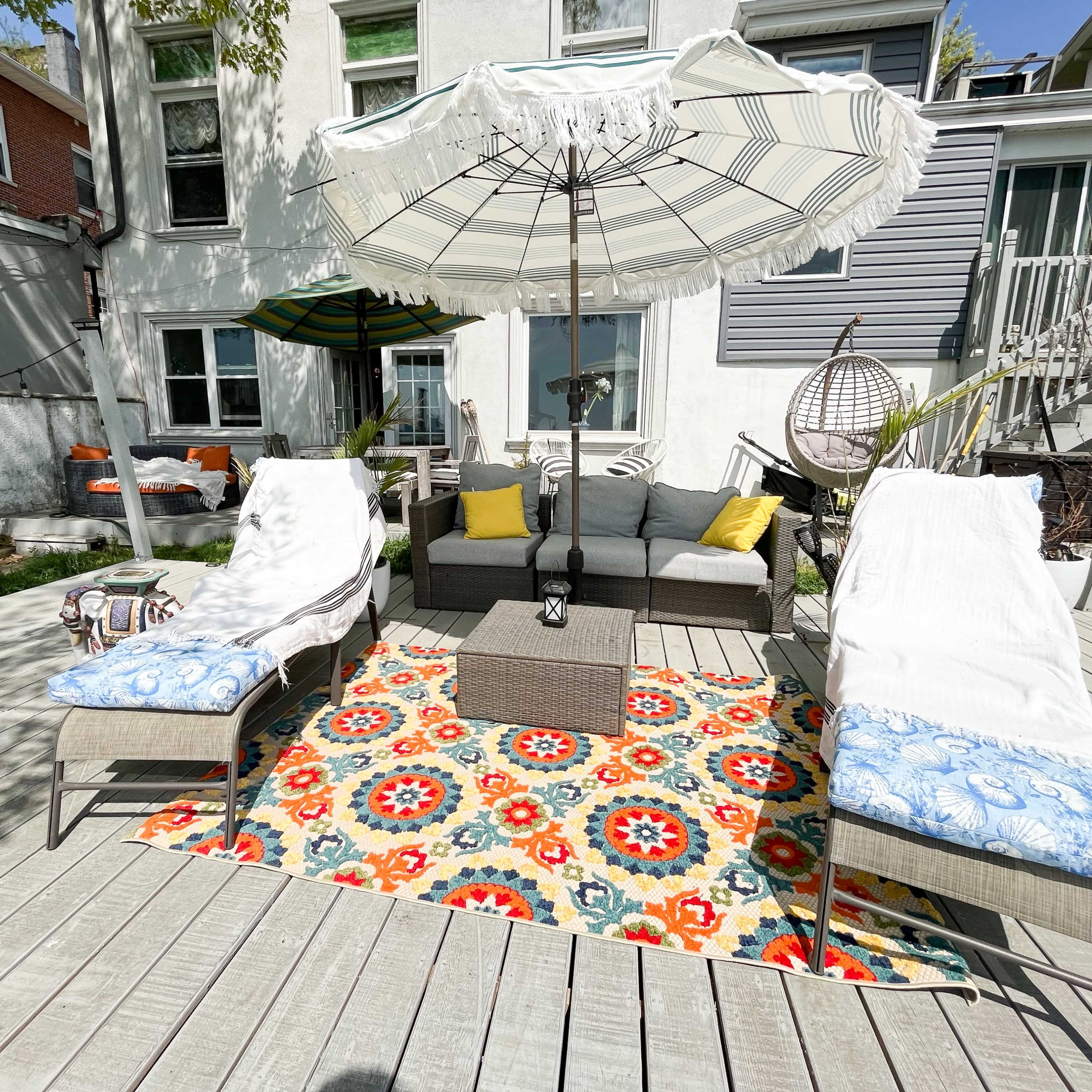 A patio area with two lounge chairs, a table, an umbrella, and a colorful rug surrounded by a house and greenery.