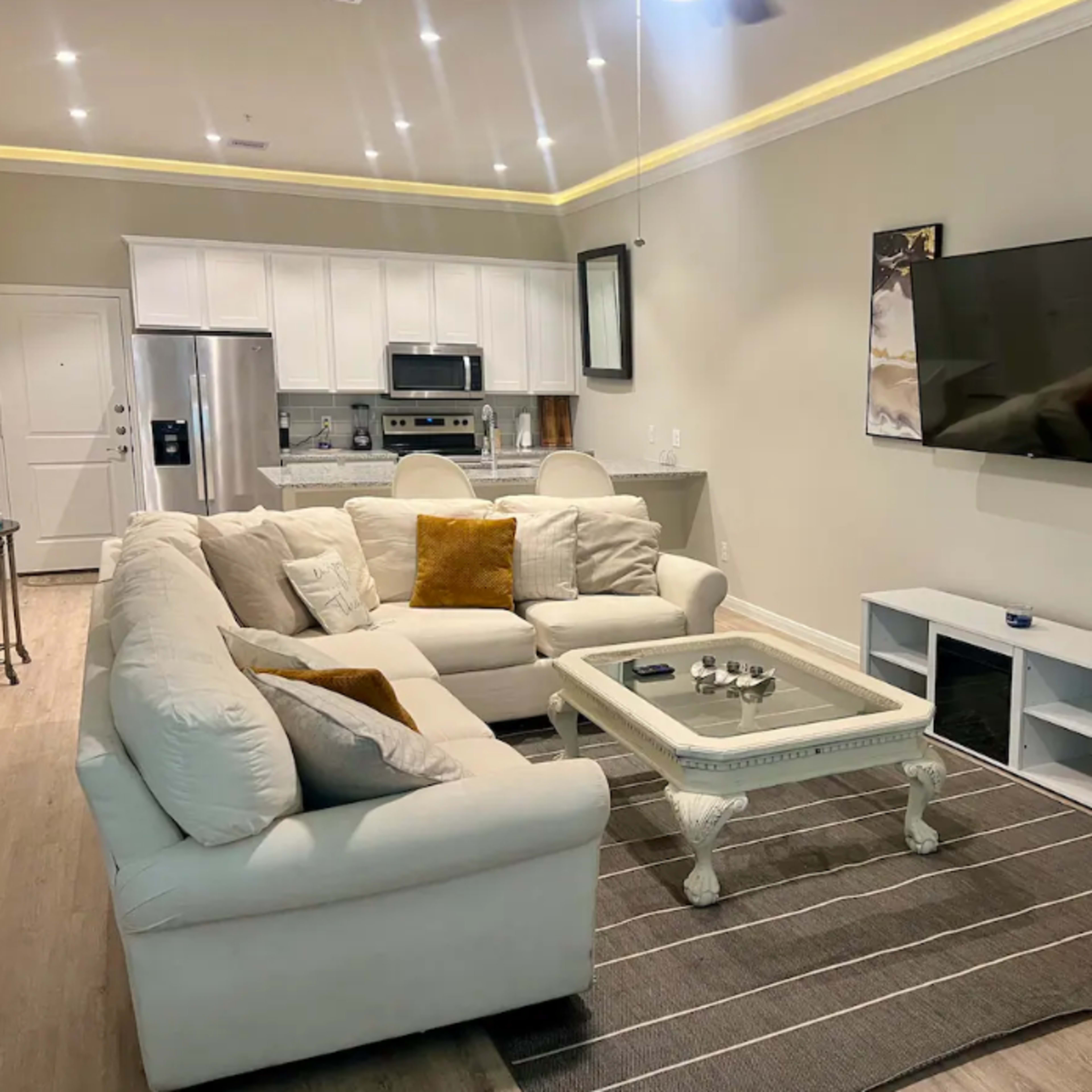 A living room with a beige sectional sofa, a white coffee table, a wall-mounted TV, and abstract artwork on the walls.