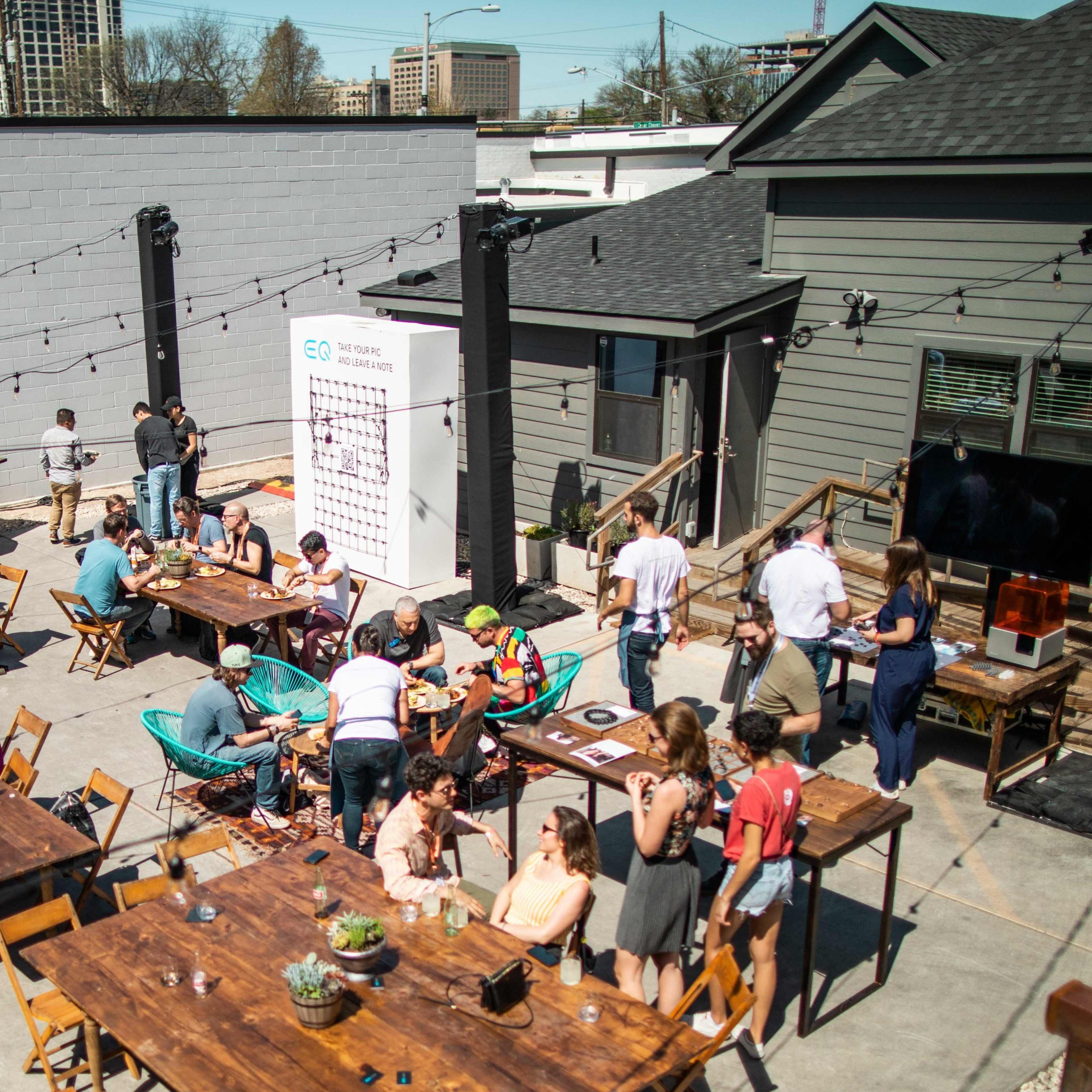A busy outdoor gathering takes place in a courtyard featuring several tables, string lights, and people enjoying food and drinks.