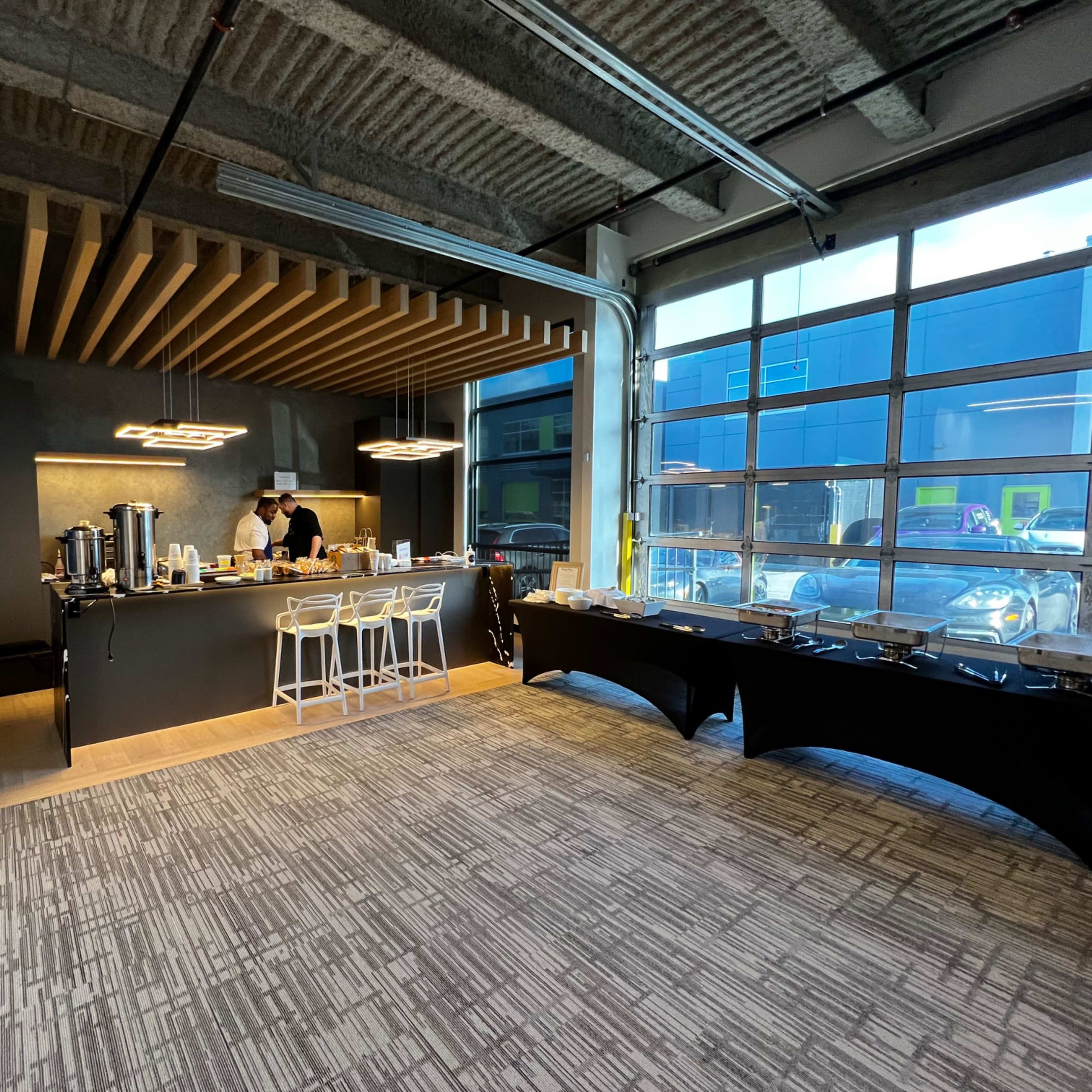 A modern indoor space features a dark countertop bar with a person preparing food, surrounded by chairs and a buffet table, with large windows allowing natural light.
