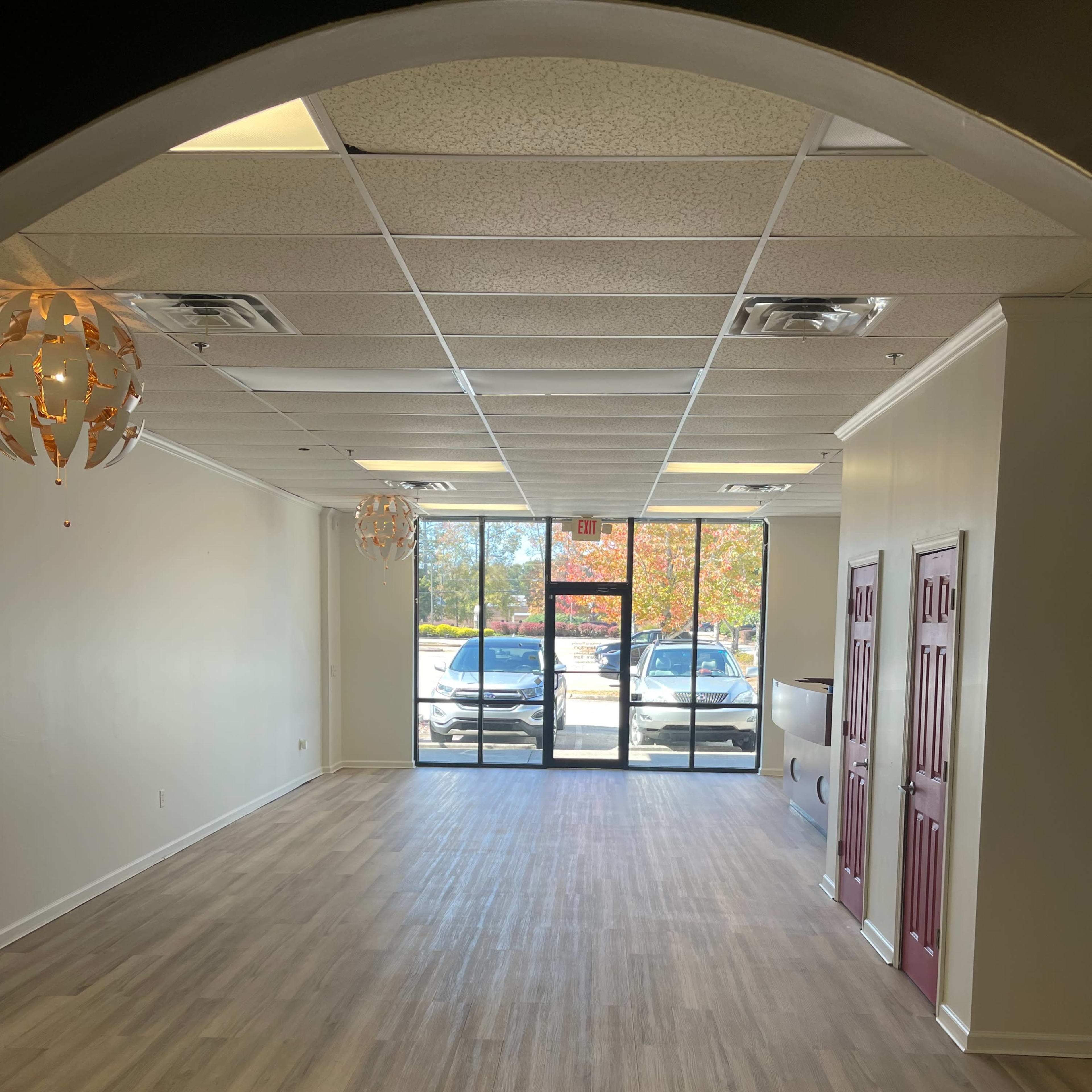 An empty commercial space with light-colored walls, wooden flooring, and large front windows allowing natural light.
