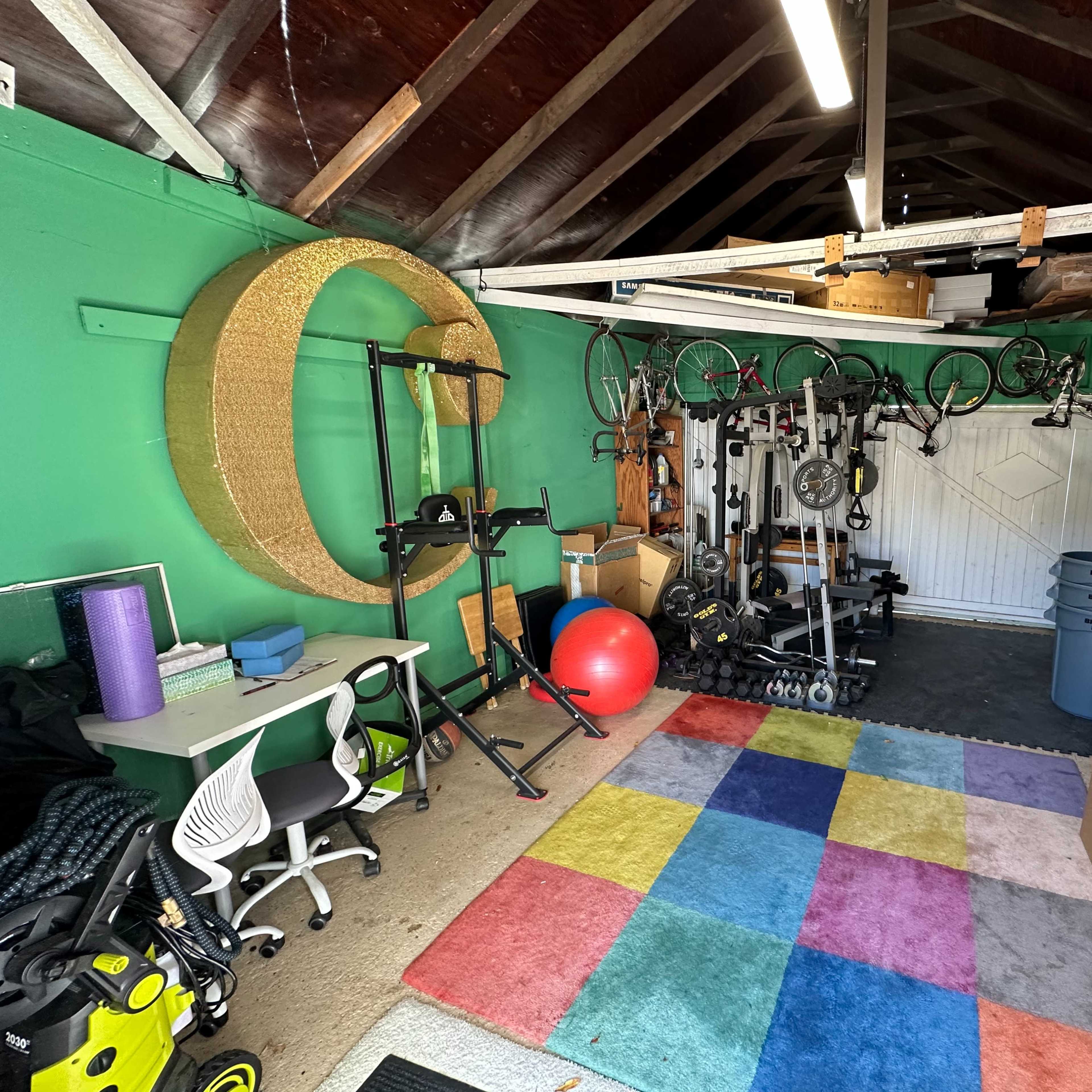 A garage space with exercise equipment, colorful flooring, and storage items, including bikes hanging from the ceiling and boxes stacked against the walls.