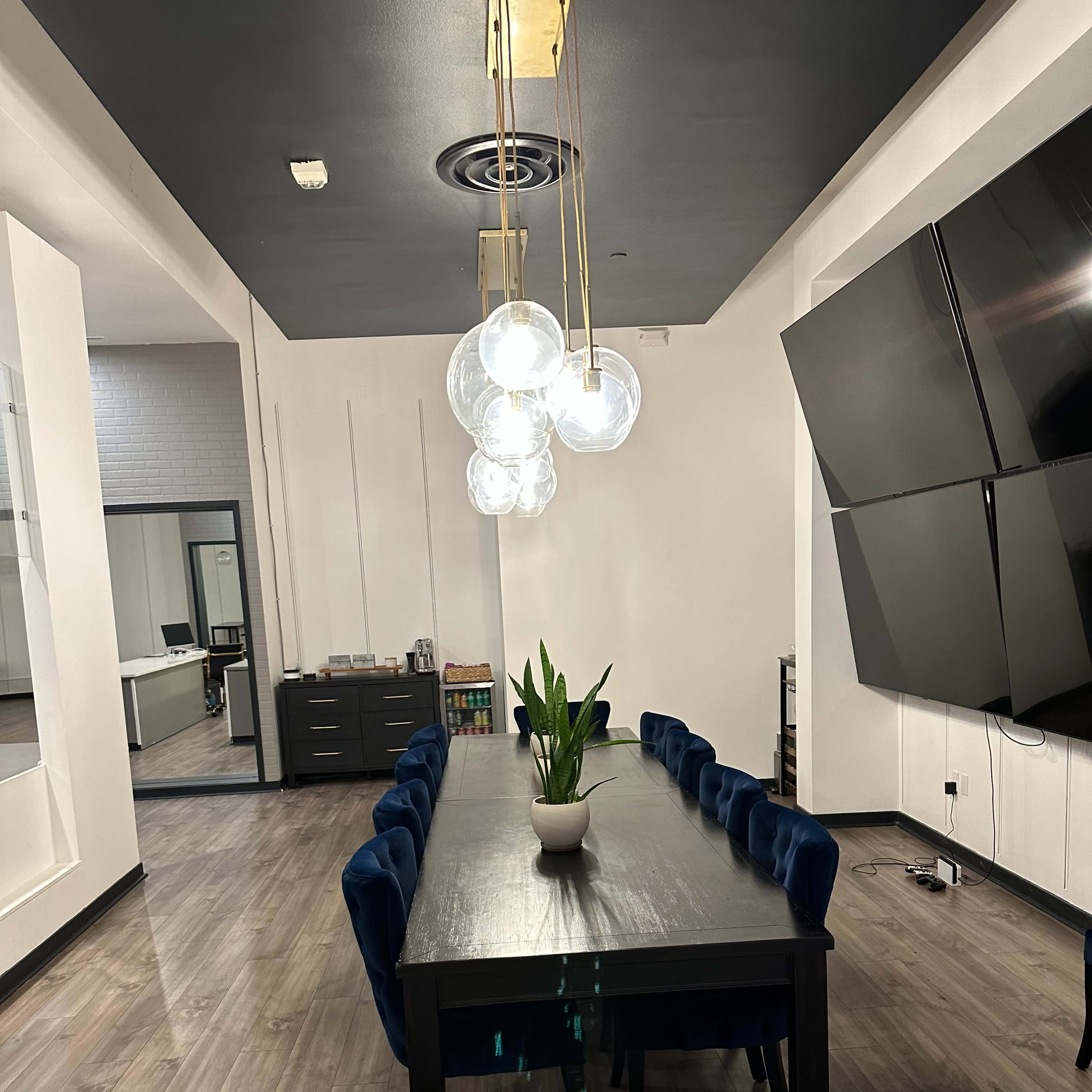 A long, dark wooden dining table with blue velvet chairs is illuminated by pendant lights in a modern room featuring a large television on one wall.