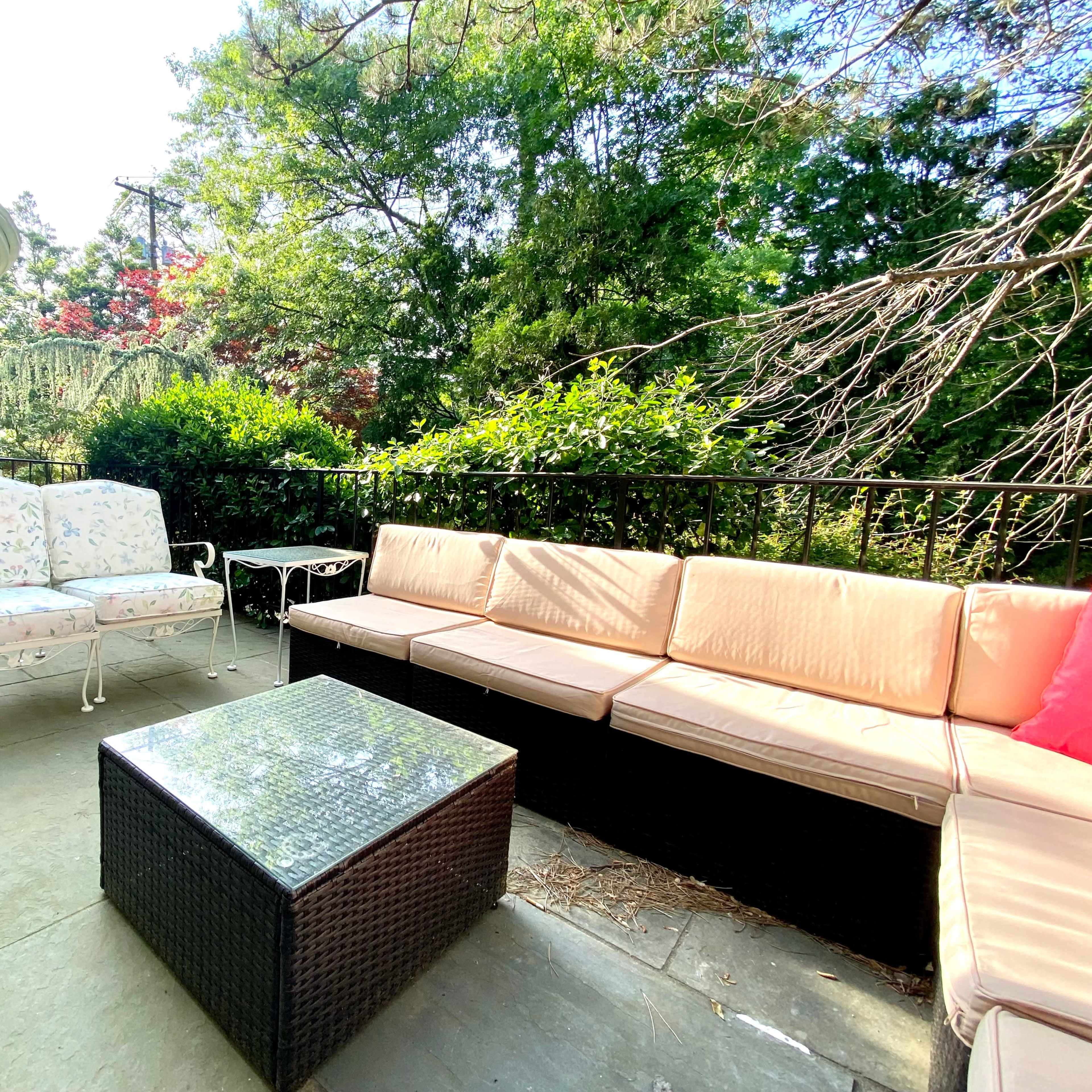 A stone patio area with a sectional couch, a small end table, and two chairs surrounded by greenery.
