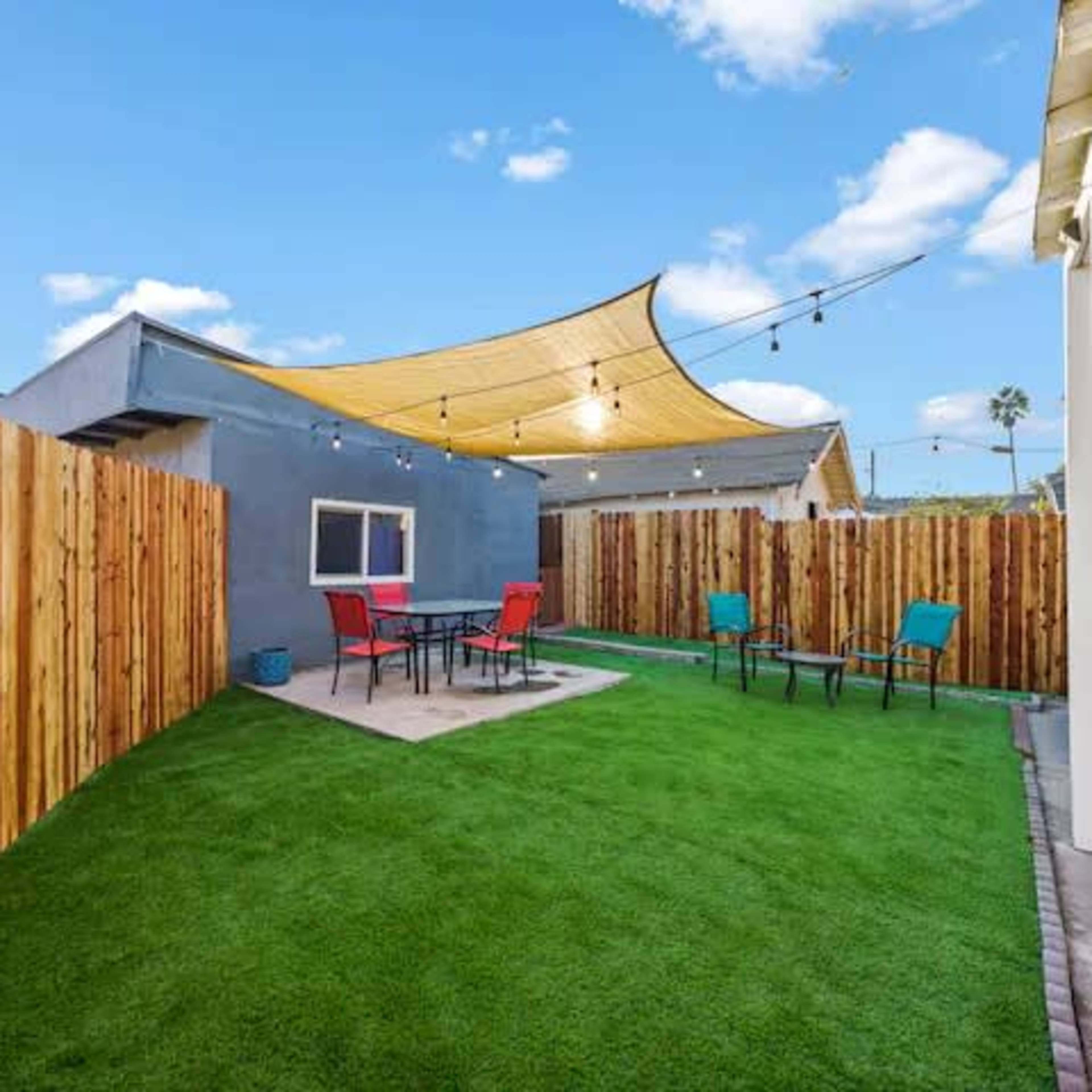 A backyard with artificial grass, a seating area with four chairs and a table, and a large shade sail overhead, surrounded by wooden fences.