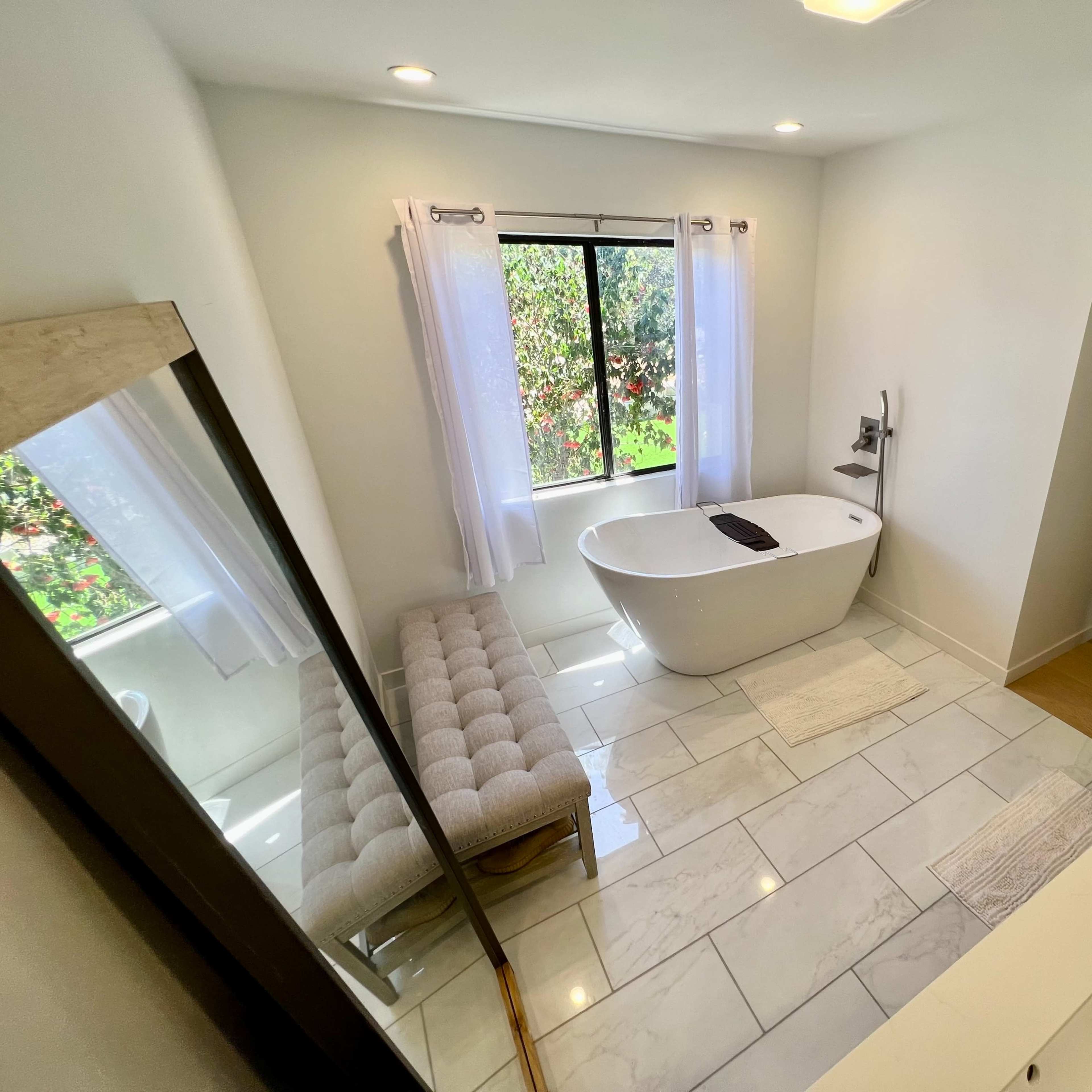 A modern bathroom featuring a freestanding bathtub, a double vanity sink, and a large window with sheer white curtains allowing natural light.