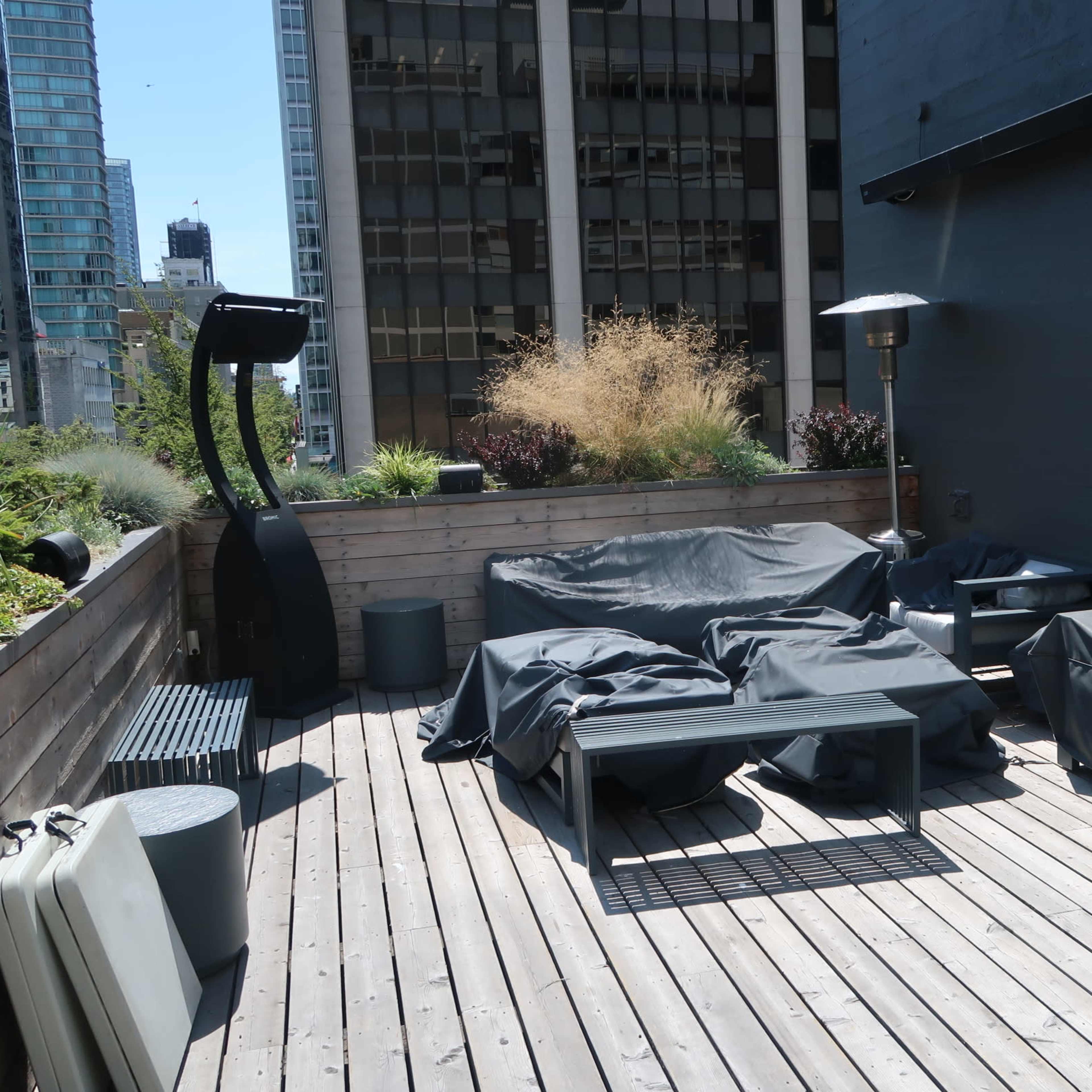 A rooftop deck features covered furniture, a heater, and surrounding plants amid tall buildings.