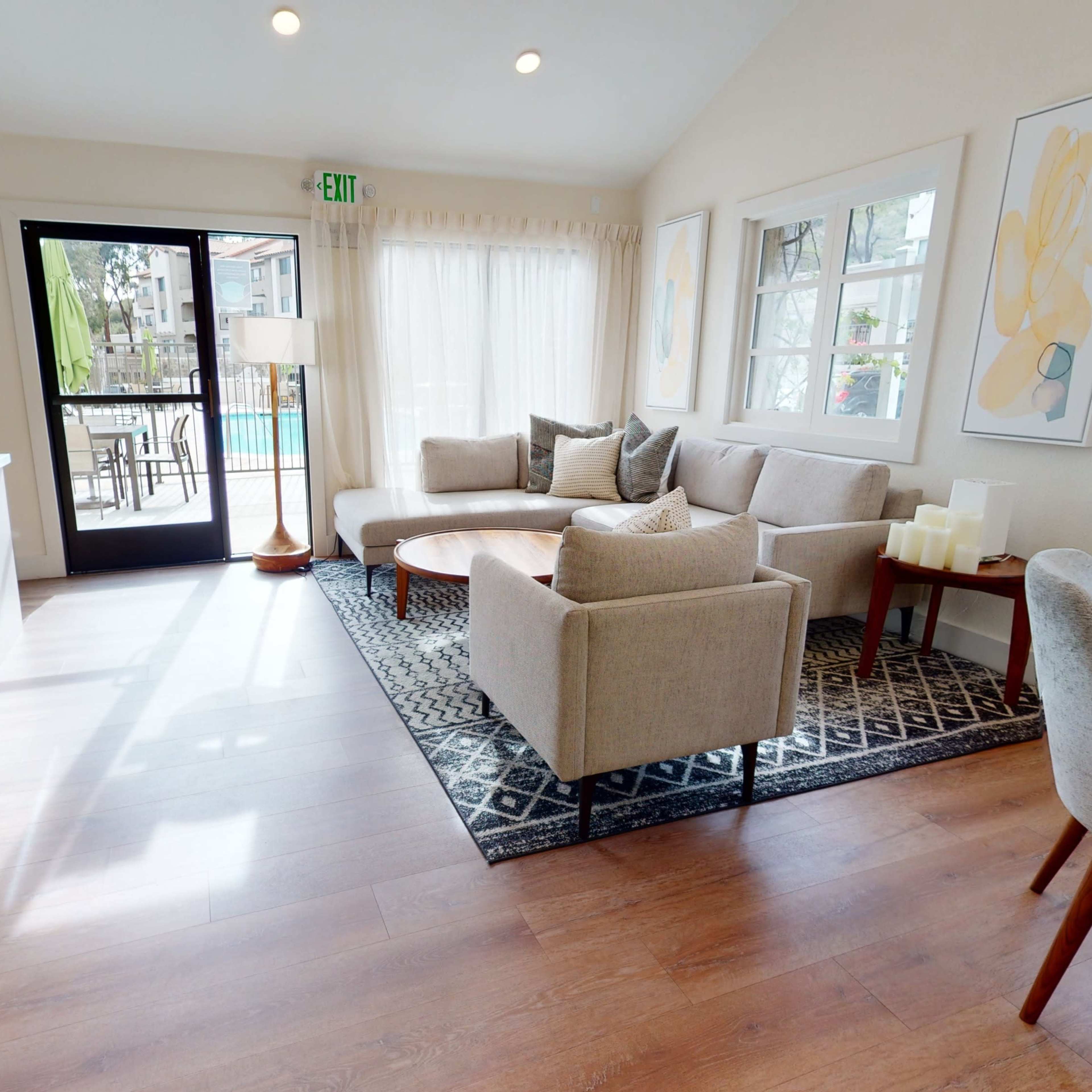 A sunlit living room featuring a beige couch, matching chairs, a wooden table, and large windows overlooking a pool area.
